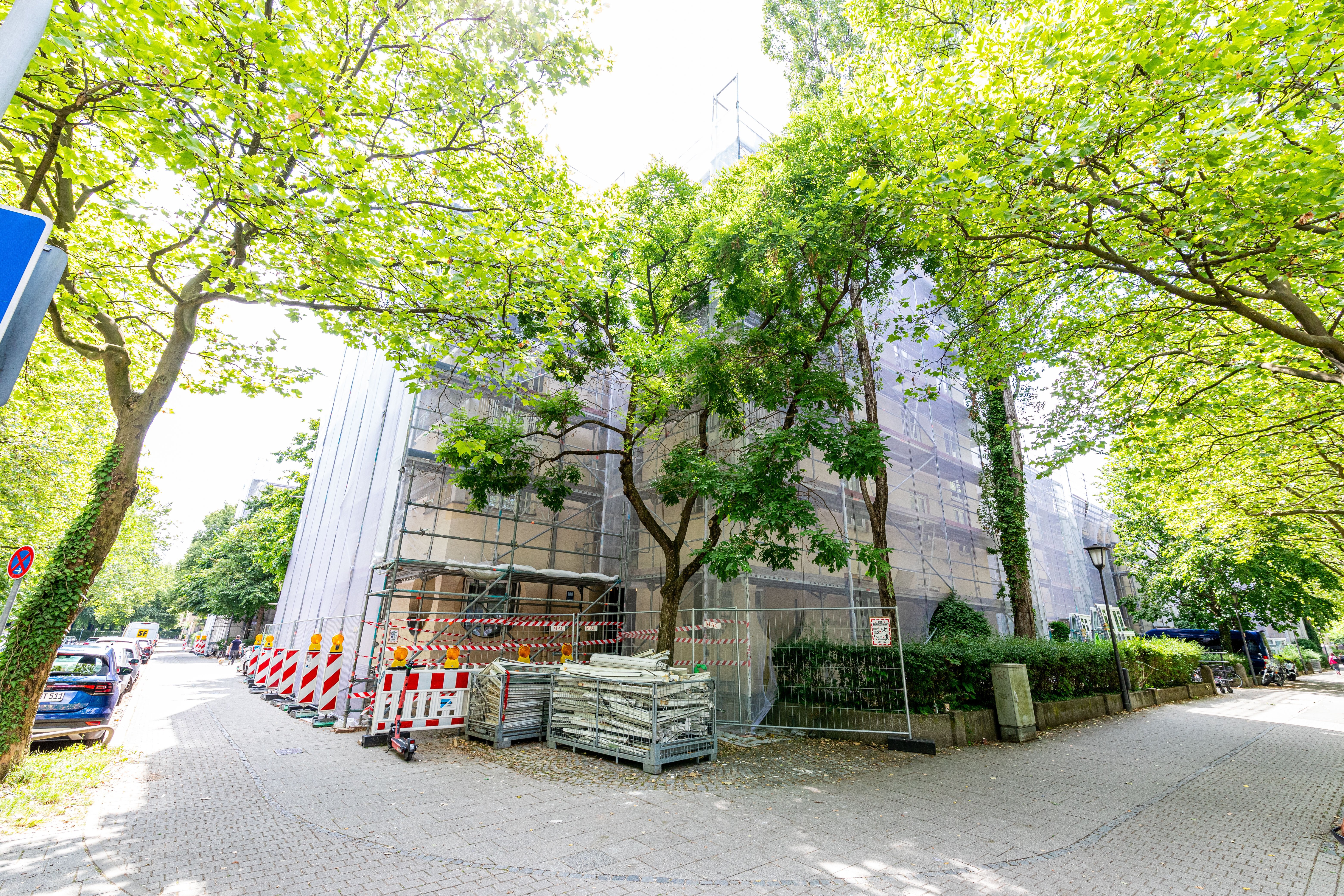 Ein Wohngebäude ist von Gerüsten und weißen Planen umgeben, mit Bäumen im Vordergrund und Hintergrund. Absperrungen und Baumaterialien befinden sich auf dem Gehweg. Eine Straße mit geparkten Autos ist links zu sehen.