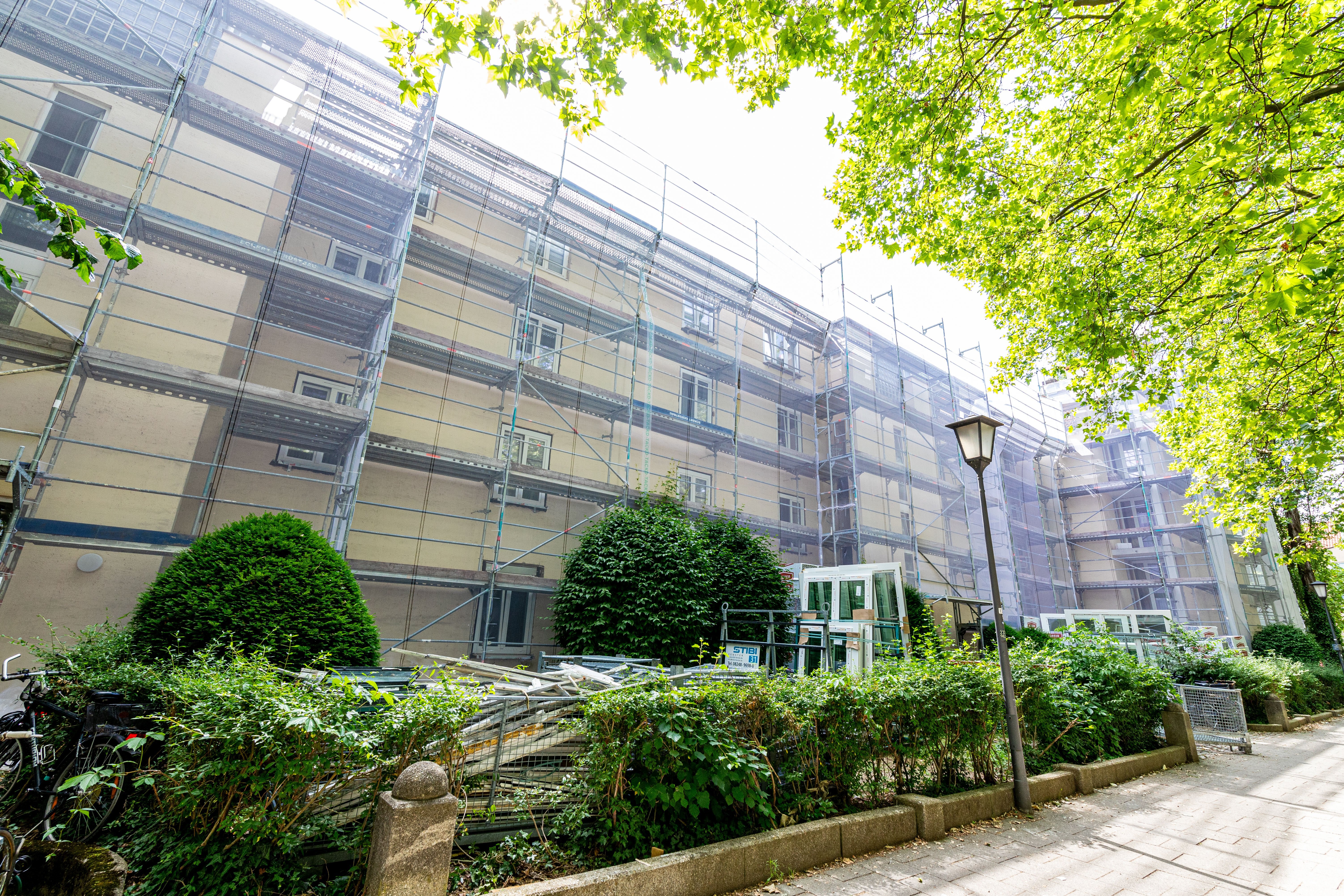  Ein mehrstöckiges Wohngebäude ist vollständig mit Baugerüsten bedeckt. Im Vordergrund sind grüne Büsche und Bäume zu sehen, die teilweise die Sicht auf die untere Fassade verdecken. Ein Laternenpfahl steht am rechten Bildrand, und ein Gehweg verläuft parallel zum Gebäude.