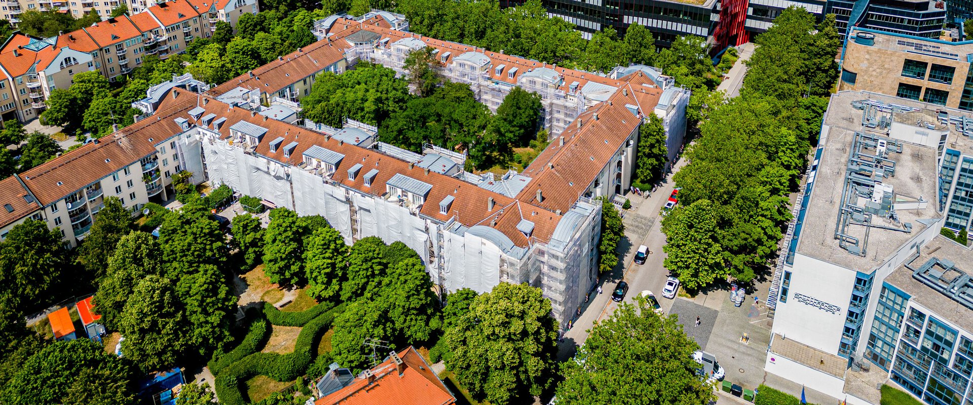 Luftaufnahme des Berliner Quartiers in München. Eine große U-förmige Wohnanlage ist von Bäumen umgeben. Teile der Gebäudefassaden sind mit weißem Material bedeckt. Im Hintergrund sind weitere Stadtgebäude und ein hoher Wolkenkratzer zu sehen.