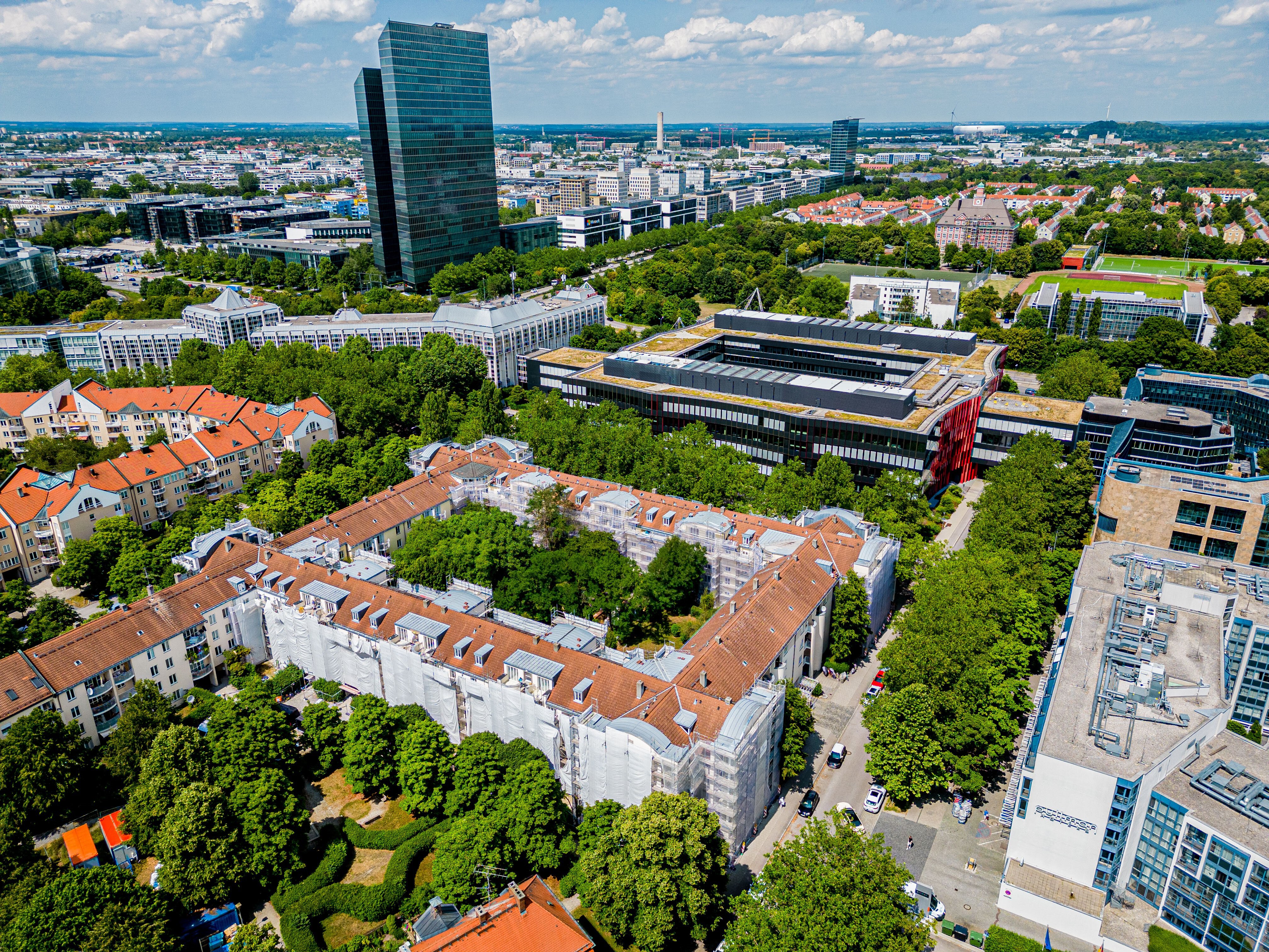 Luftaufnahme des Berliner Quartiers in München. Eine große U-förmige Wohnanlage ist von Bäumen umgeben. Teile der Gebäudefassaden sind mit weißem Material bedeckt. Im Hintergrund sind weitere Stadtgebäude und ein hoher Wolkenkratzer zu sehen.