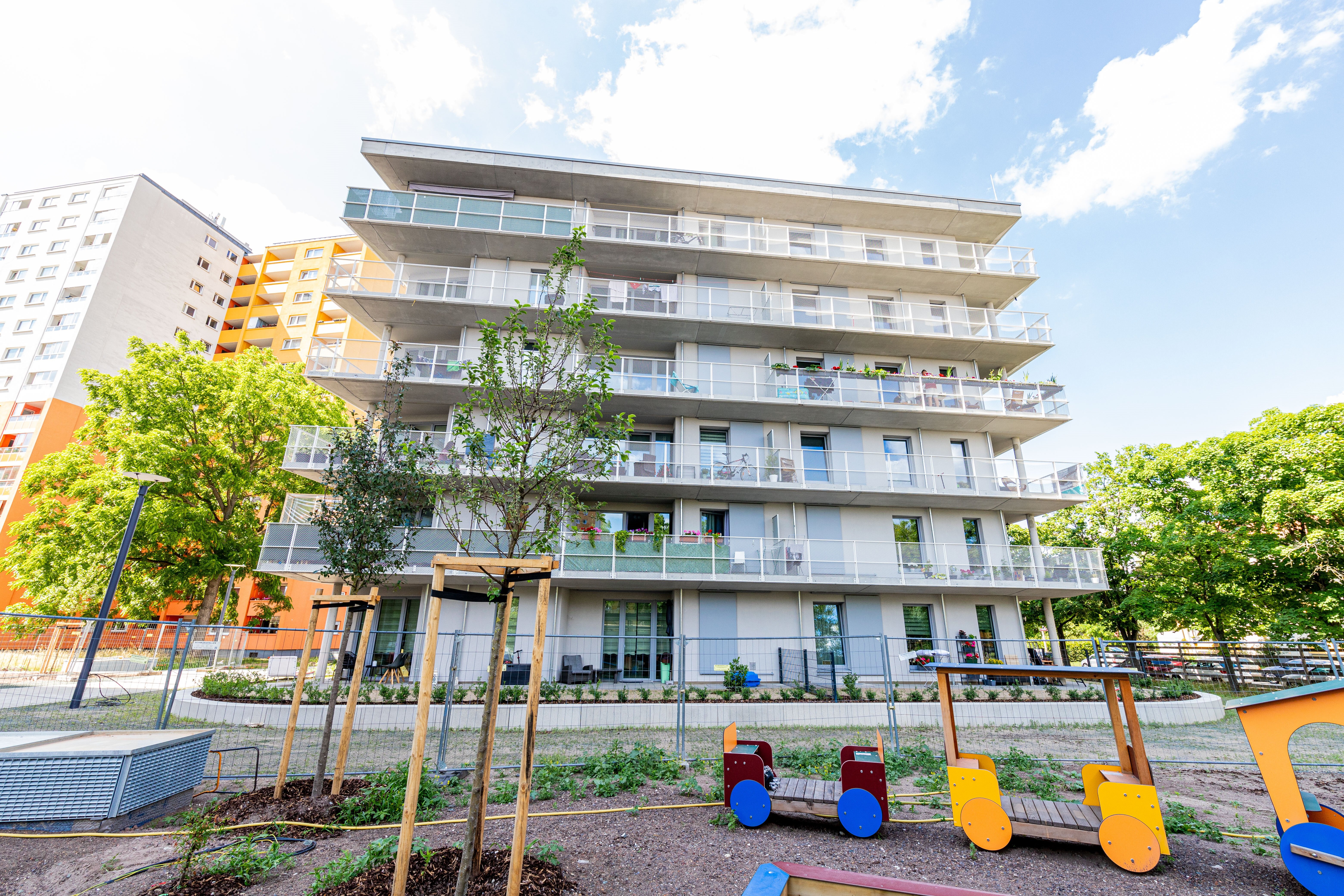 Modernes Mehrfamilienhaus mit Balkonen, davor ein Spielplatzbereich mit jungen Bäumen und Absperrungen. Im Hintergrund sind weitere Gebäude und Bäume sichtbar.