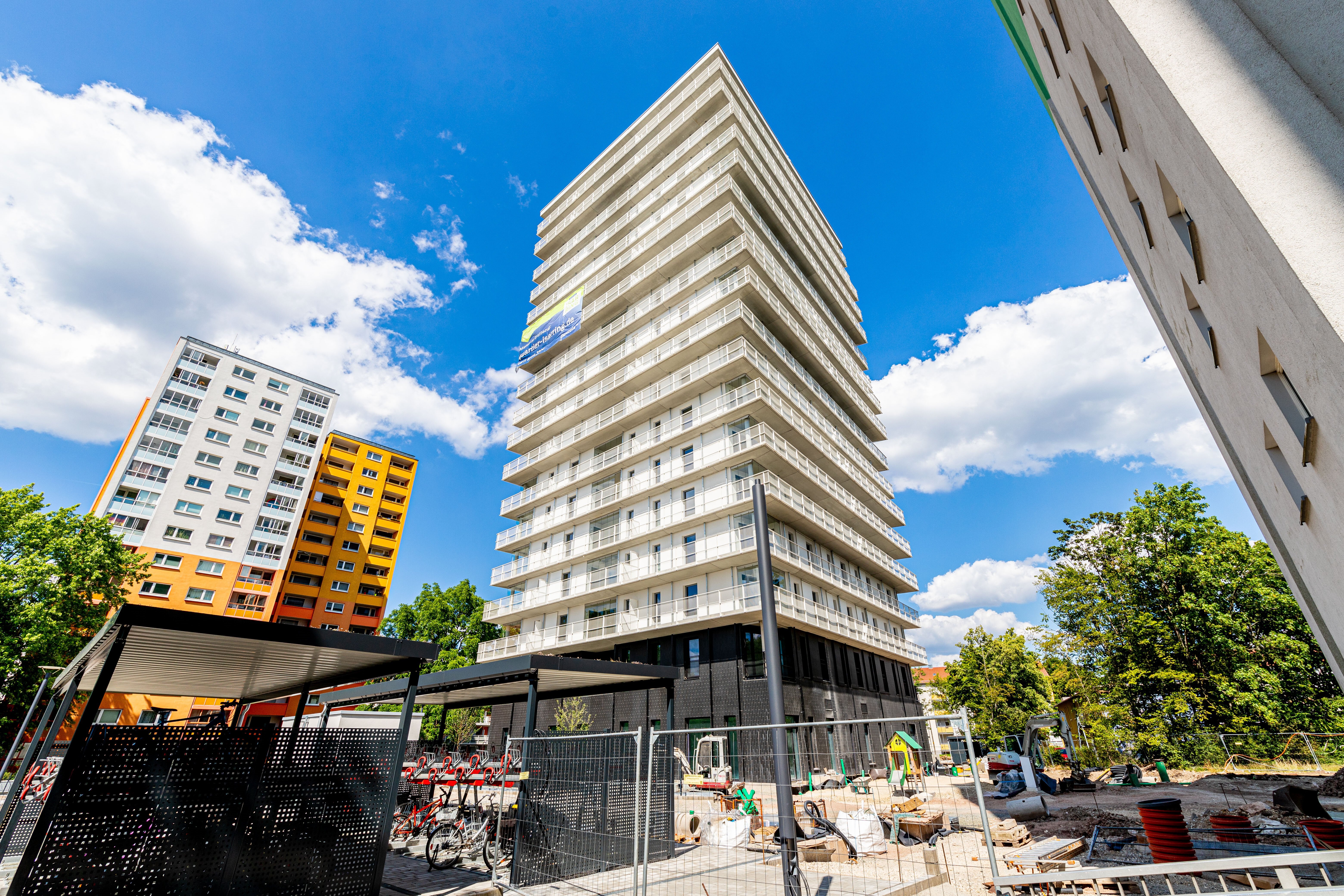 Ein hohes, modernes, weißes Gebäude mit vielen Balkonen steht prominent in der Bildmitte. Links davon ist ein weiteres Gebäude mit orangefarbenen und weißen Abschnitten teilweise sichtbar. Im Vordergrund befindet sich eine Baustelle mit Zäunen, einigen Geräten und einem überdachten Bereich mit Fahrradständern.