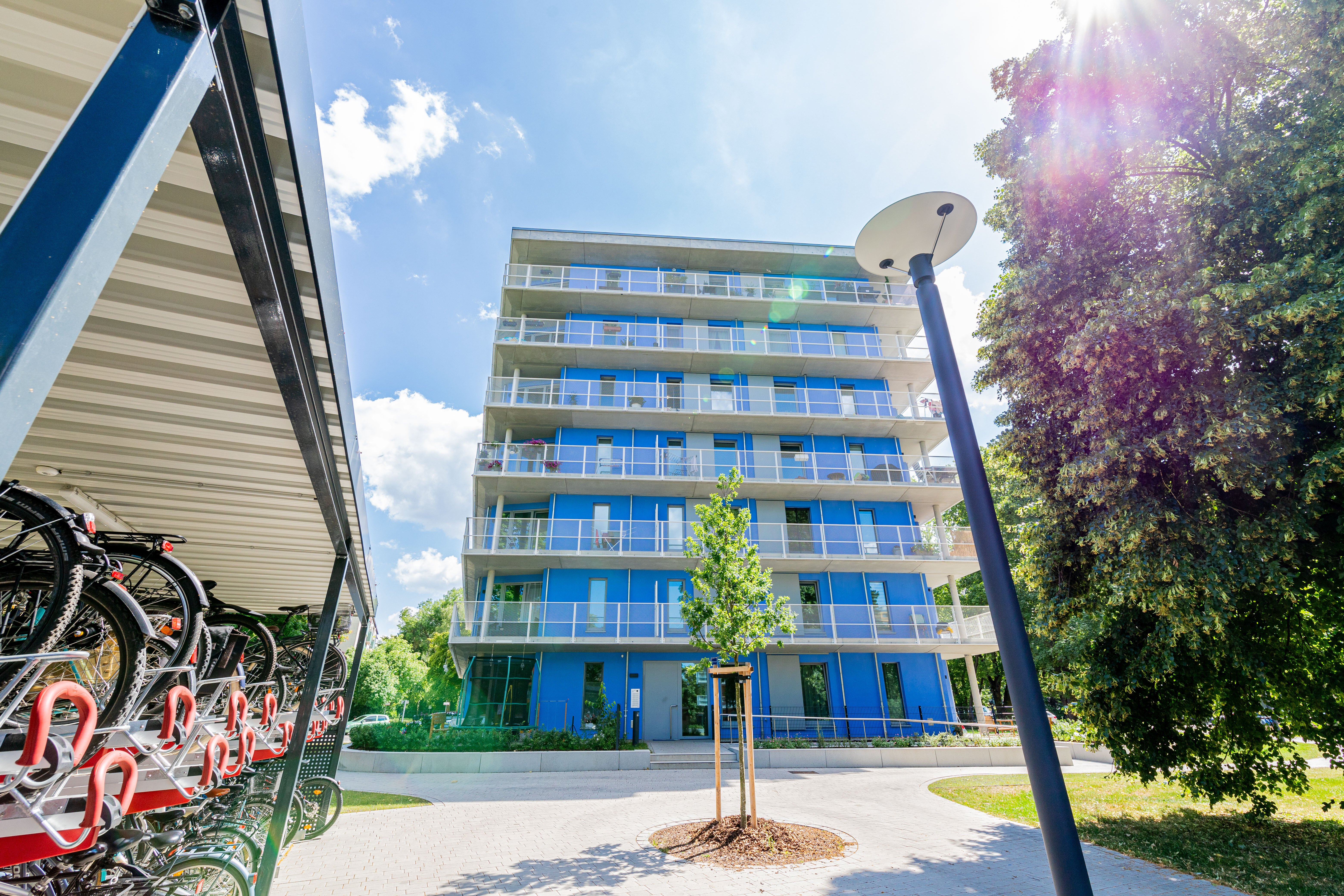 Ein modernes, mehrstöckiges Wohngebäude mit blauen und weißen Balkonen steht unter einem sonnigen Himmel. Links im Vordergrund befindet sich ein überdachter Fahrradabstellplatz mit vielen Fahrrädern. Rechts steht ein hoher, runder Laternenpfahl, und Bäume sind auf beiden Seiten des Gebäudes zu sehen. Ein gepflasterter Weg führt zum Eingang des Gebäudes.