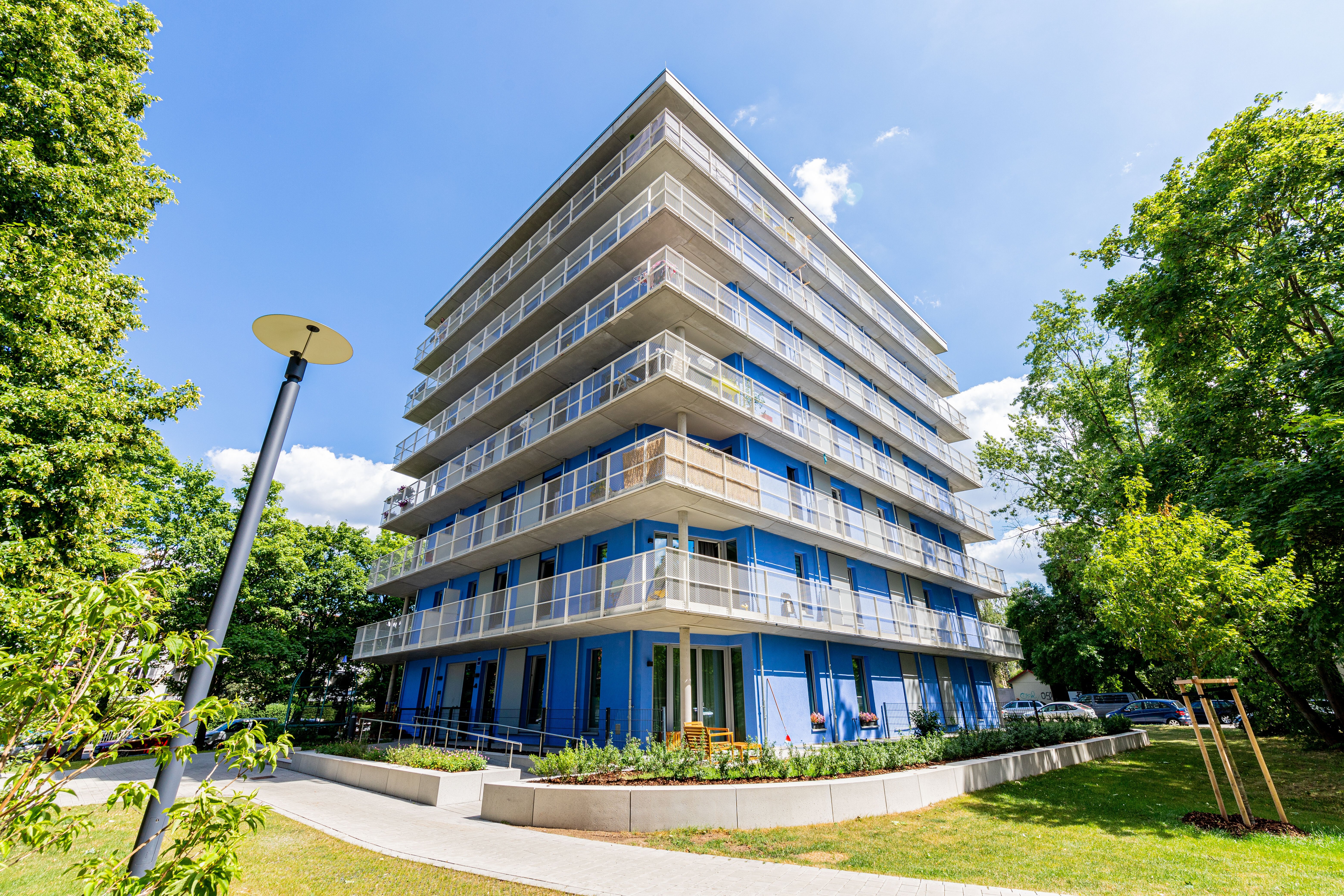 Ein modernes, mehrstöckiges Wohngebäude mit blauen Fassadenelementen und Balkonen. Es ist umgeben von grünen Bäumen und Rasenflächen. Im Vordergrund befindet sich ein gepflasterter Weg, der um das Gebäude herumführt, und ein runder Laternenpfahl steht links im Bild.