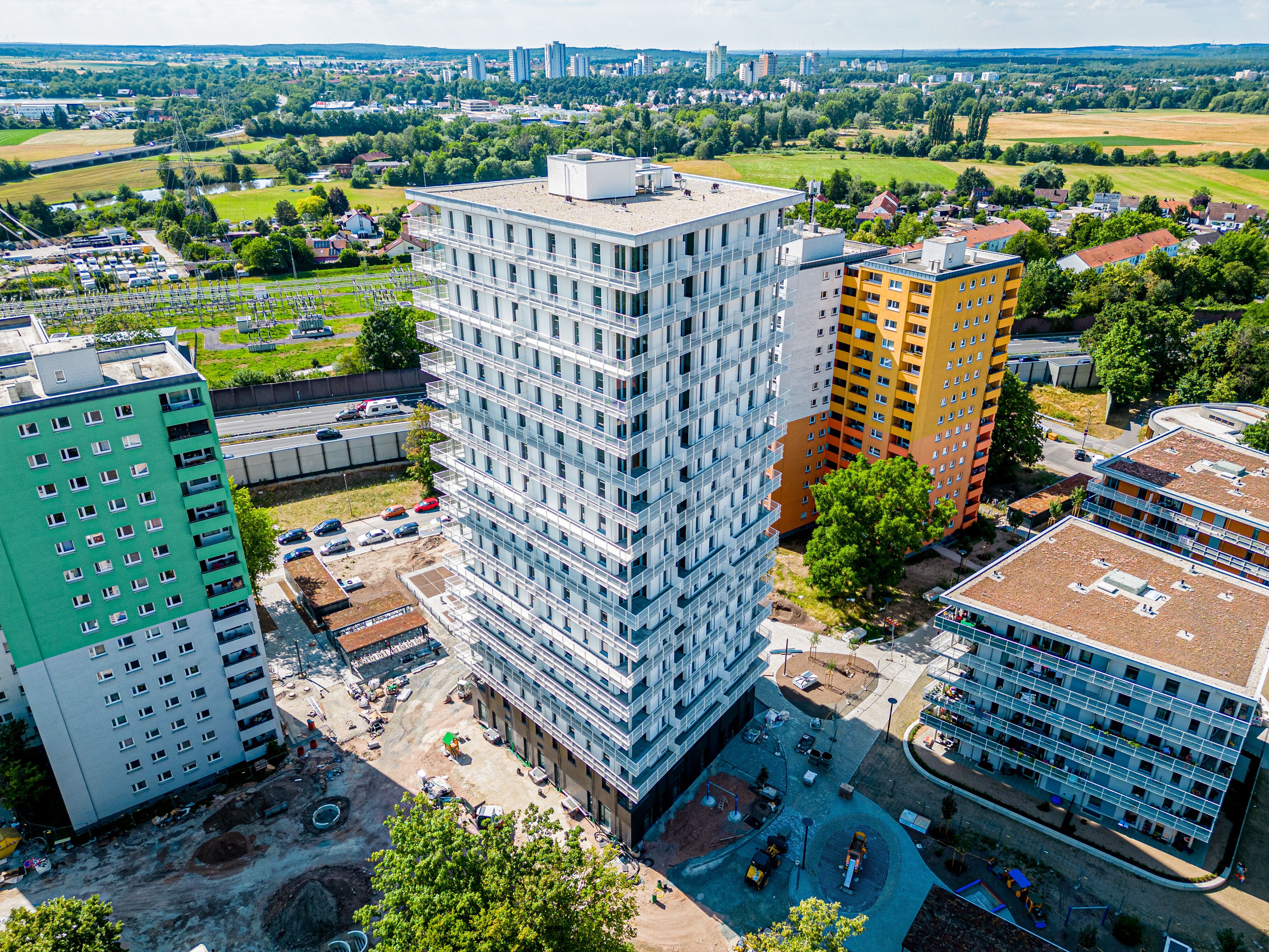 Luftaufnahme des "Quartier Isarring" in Erlangen, mit einem hohen, modernen Wohngebäude in der Mitte und weiteren, farblich unterschiedlichen Wohngebäuden daneben. Baustellenbereiche sind im Vordergrund erkennbar. Im Hintergrund erstrecken sich grüne Flächen, eine Autobahn und die Stadtsilhouette. 
