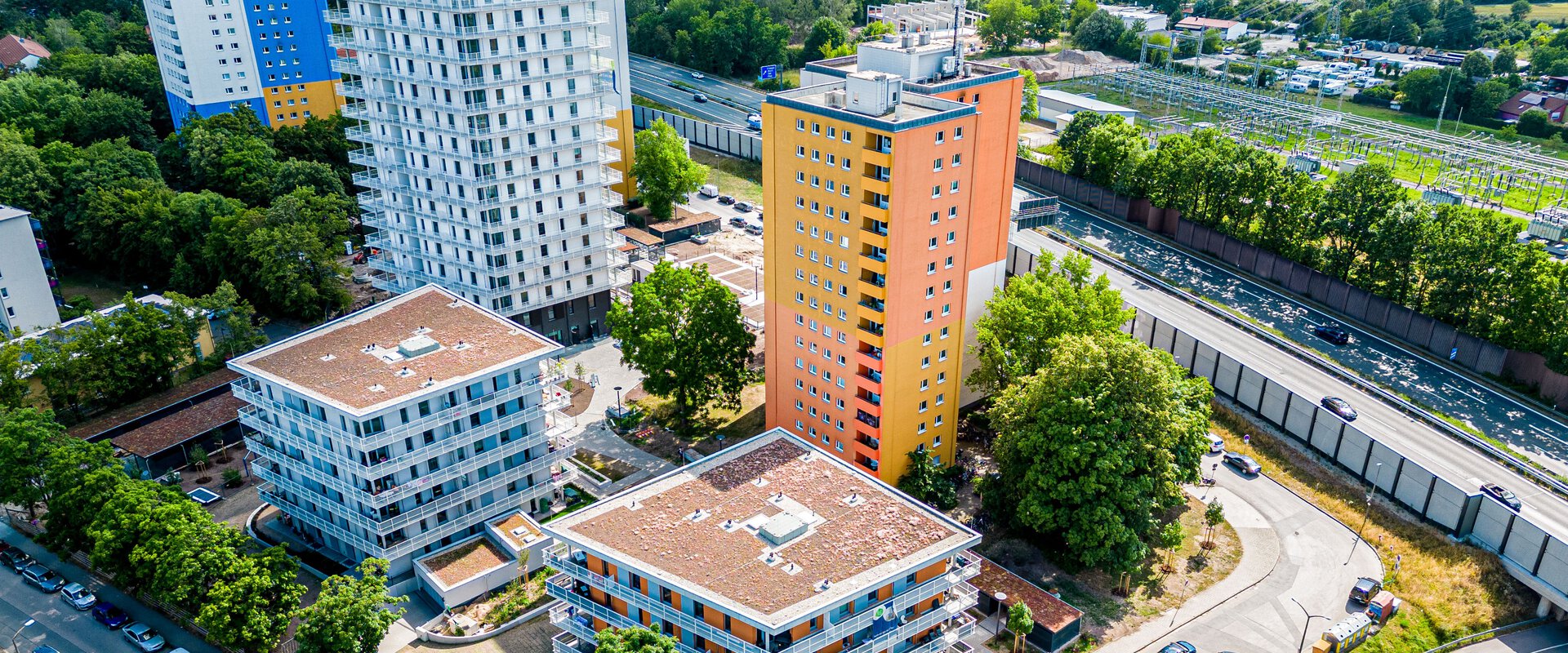 Luftaufnahme des "Quartier Isarring" in Erlangen, mit mehreren modernen Wohngebäuden unterschiedlicher Höhe und Farbe. Im Vordergrund sind vier niedrigere Gebäude zu sehen, während im Hintergrund höhere Gebäude und eine Autobahn sichtbar sind. 