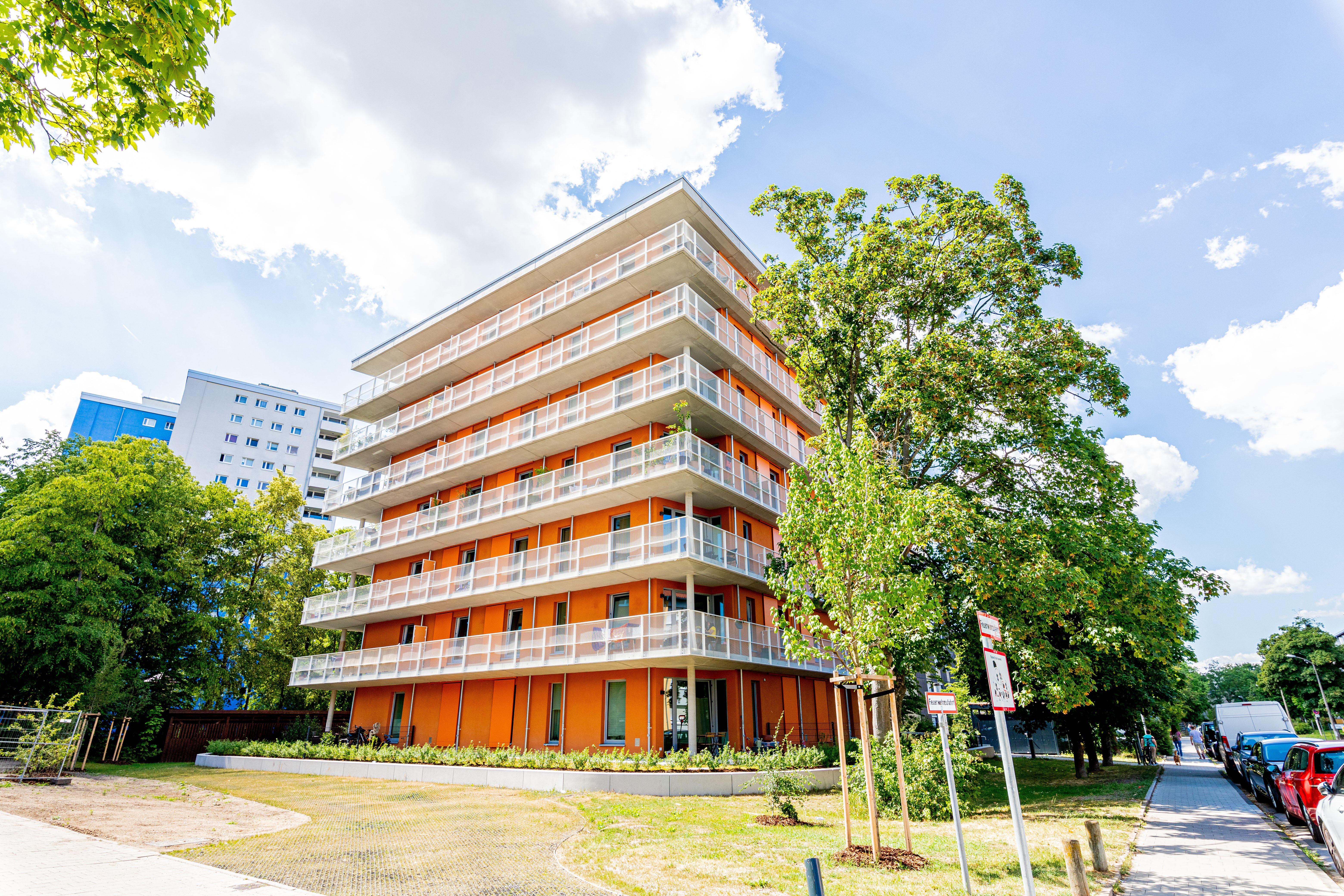 Ein modernes, mehrstöchiges Wohngebäude mit orangefarbener Fassade und weißen Balkonen. Im Hintergrund sind weitere Gebäude, darunter ein höheres blau-weißes Hochhaus, sowie grüne Bäume und eine Straße mit geparkten Autos zu sehen. Im Vordergrund erstreckt sich eine gepflegte Grünfläche.