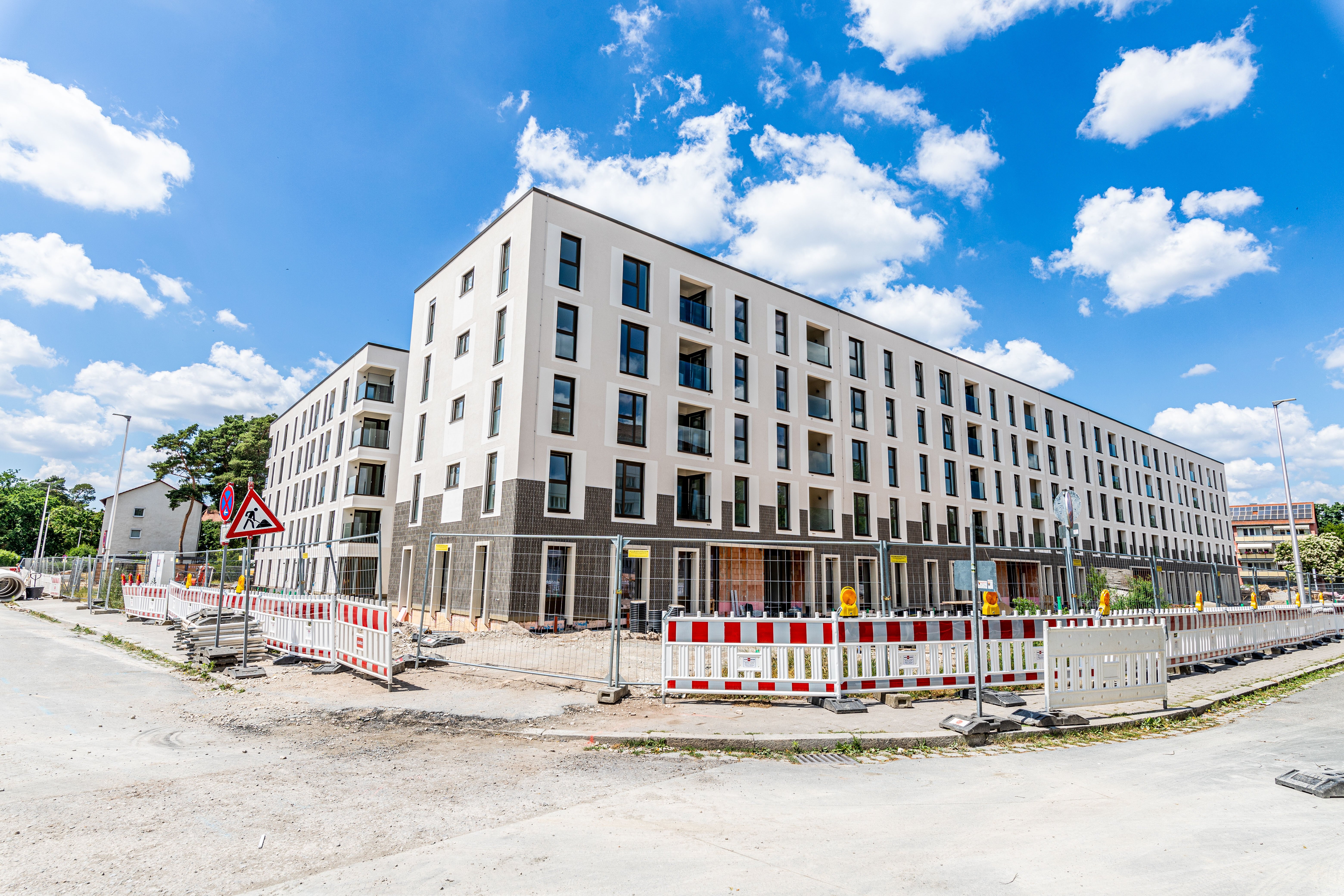 Ein langes, modernes, weißes Wohngebäude mit dunklen Fensterrahmen und einem dunklen Sockel ist im Bau und von rot-weißen Bauzäunen umgeben. Links und rechts führen Straßen entlang.