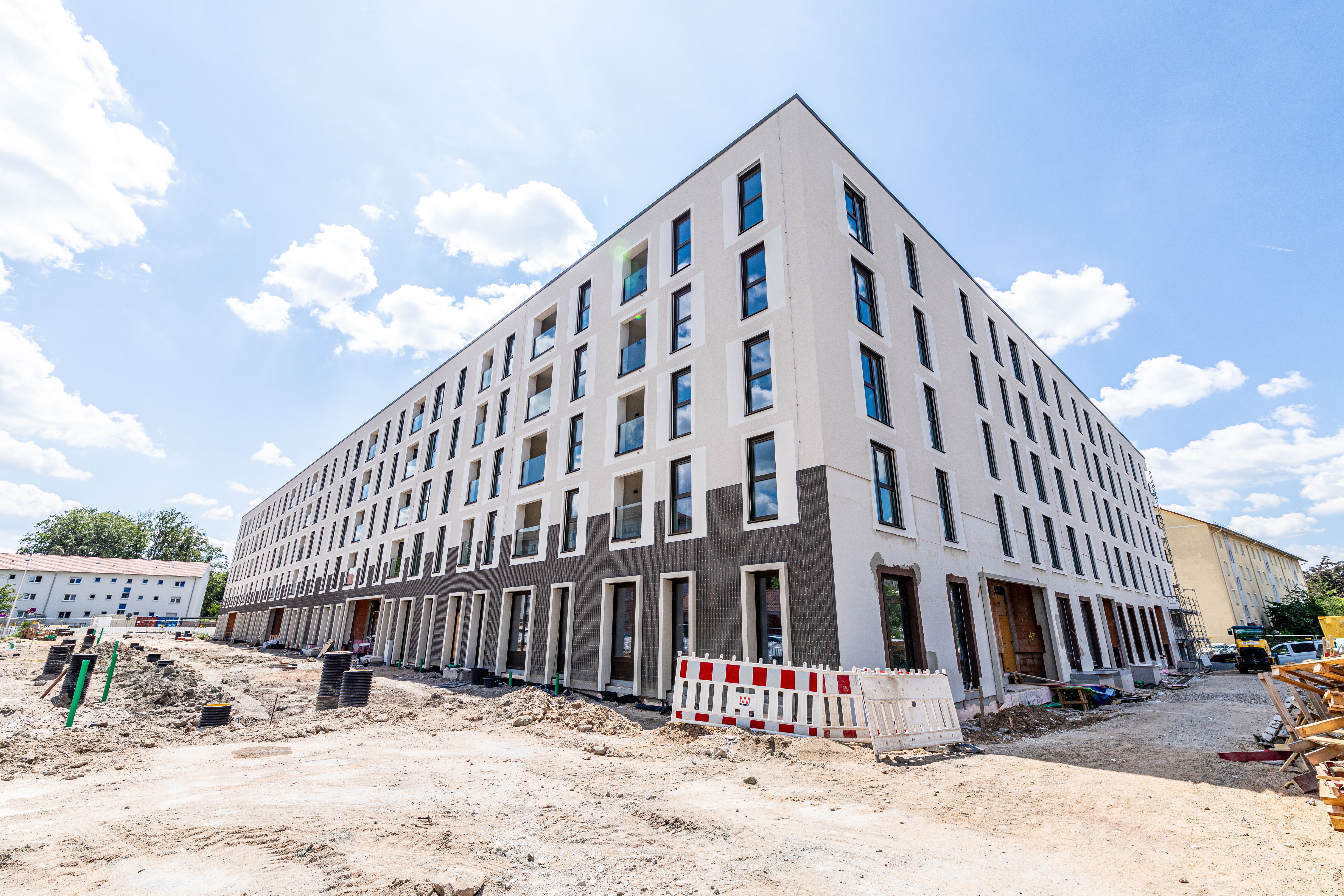  Ein langes, modernes, weißes Wohngebäude mit dunklen Fensterrahmen und einem dunklen Sockel ist im Bau und von rot-weißen Bauzäunen umgeben. Links und rechts führen Straßen entlang.