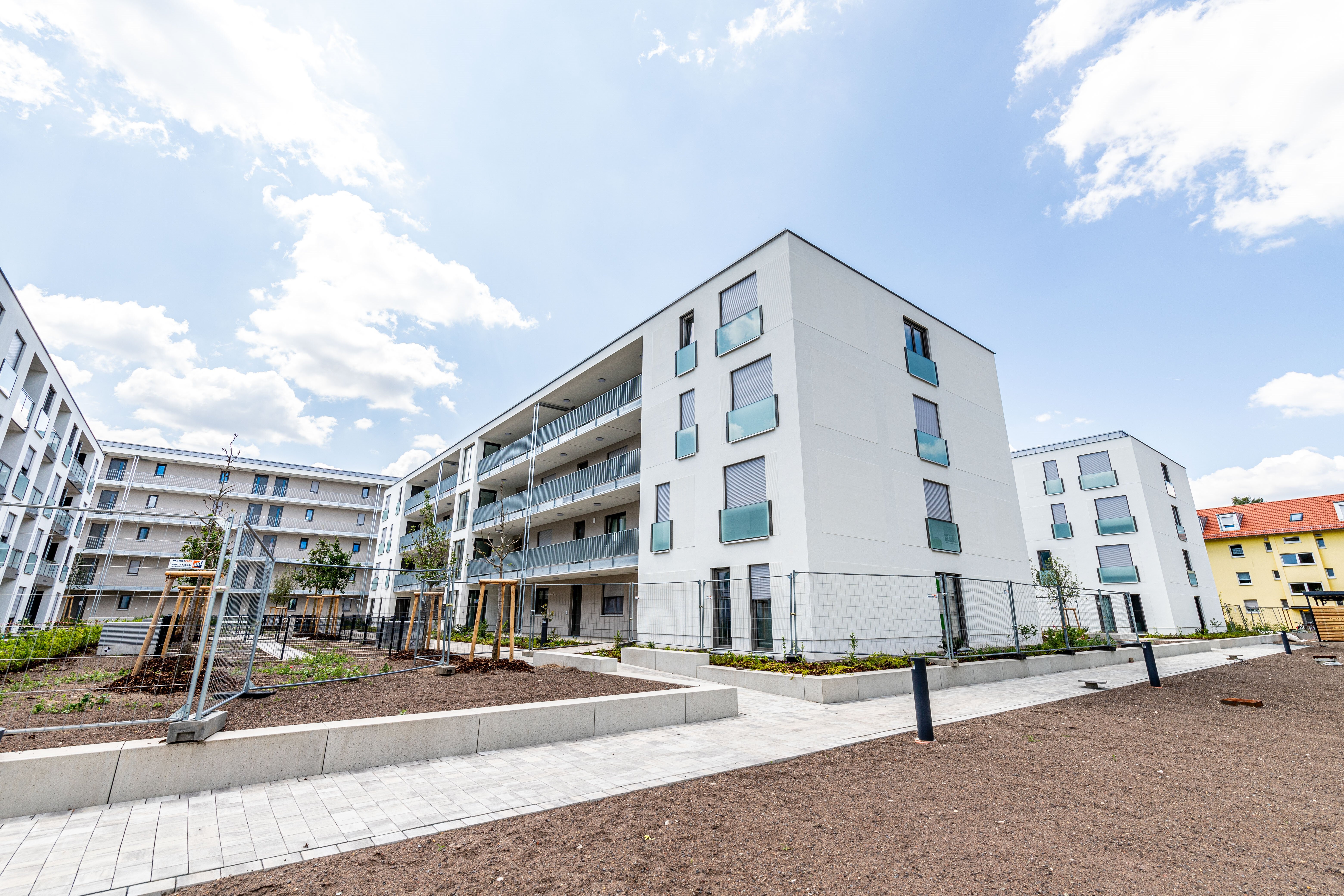 Ein moderner Gebäudekomplex mit weißen Fassaden und blauen Glasbalkonen unter einem bewölkten Himmel. Im Vordergrund befindet sich ein noch unbebauter Außenbereich mit Wegen und einigen jungen Pflanzen.