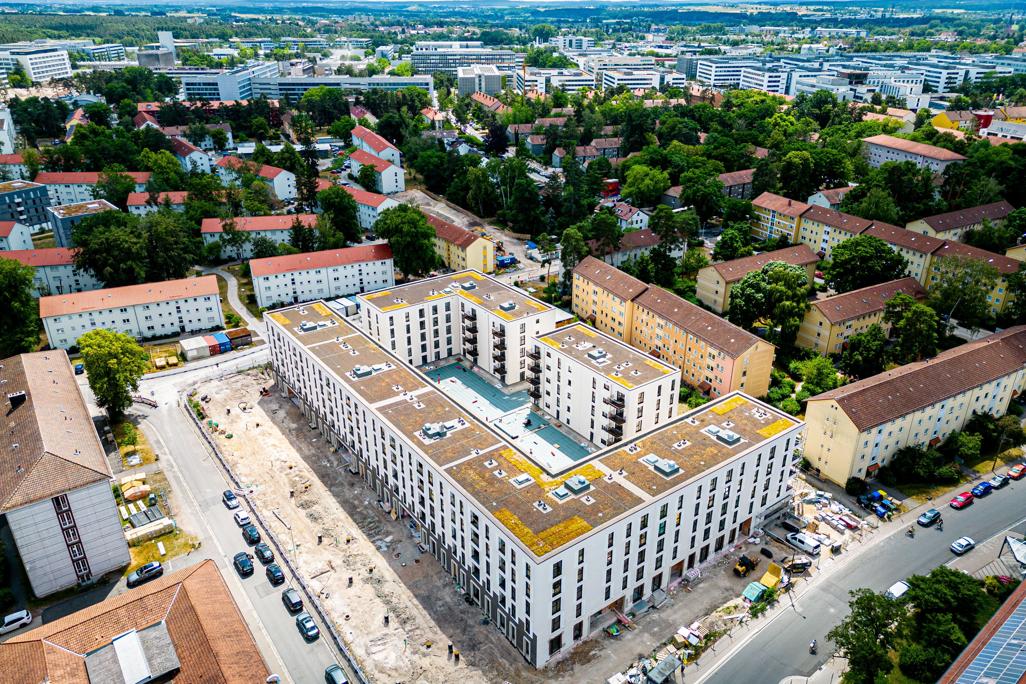  Luftaufnahme eines großen, modernen U-förmigen Wohnkomplexes mit einem Innenhof, umgeben von älteren Wohngebäuden und viel Grün. Davor und daneben befinden sich Straßen mit parkenden Autos.