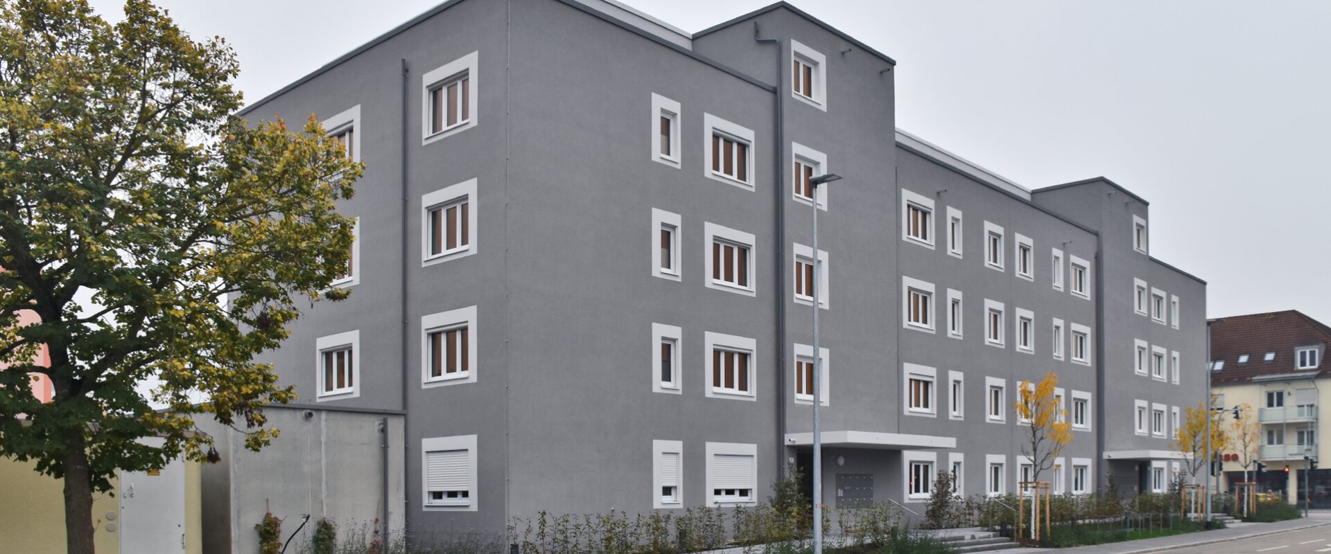 Mehrstöckiges graues Wohngebäude mit vielen Fenstern und neu gepflanzten Bäumen an der Straße