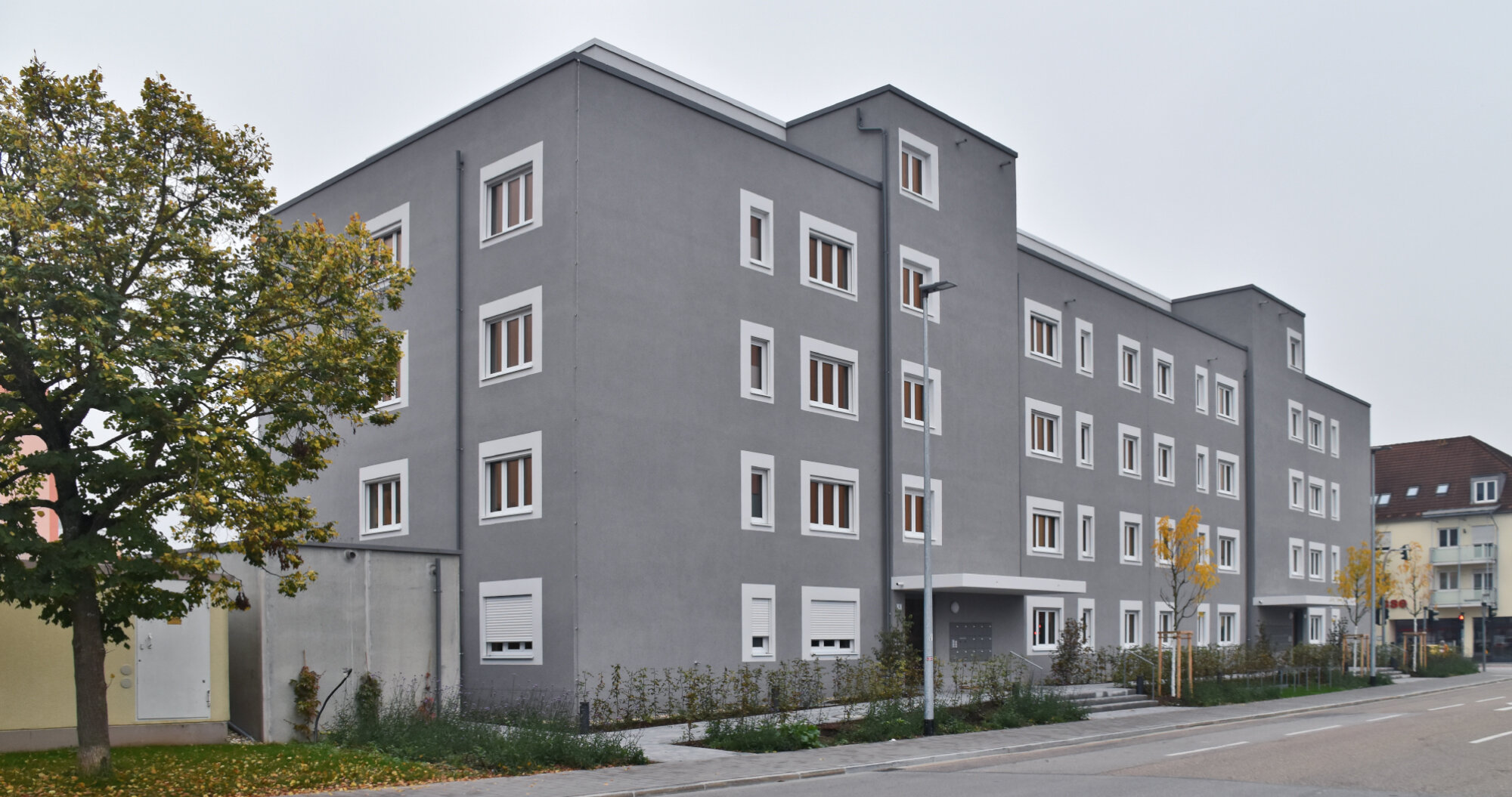 Mehrstöckiges graues Wohngebäude mit vielen Fenstern und neu gepflanzten Bäumen an der Straße