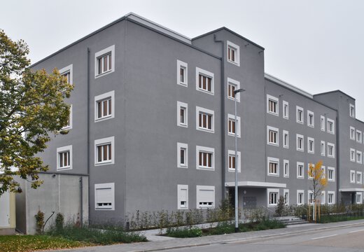 Mehrstöckiges graues Wohngebäude mit vielen Fenstern und neu gepflanzten Bäumen an der Straße