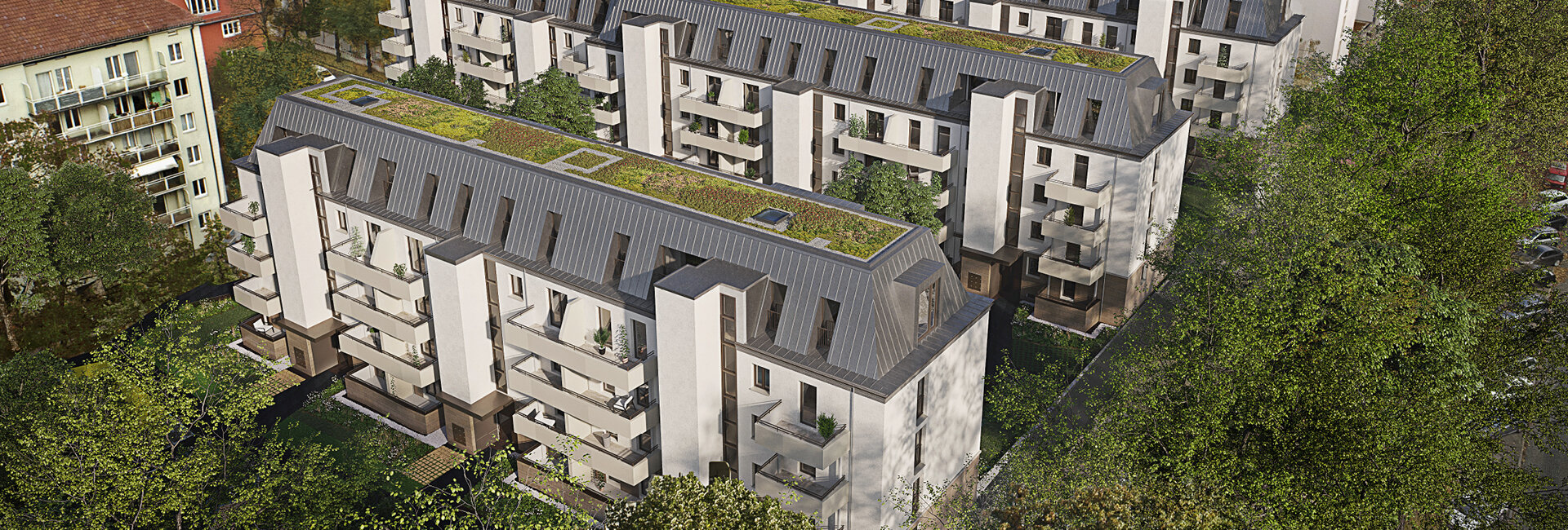 Luftaufnahme von mehreren modernen Wohngebäuden mit dunkelgrauen Mansarddächern, die teilweise begrünt sind. Die Fassaden sind hell mit hellgrauen Balkonen. Die Gebäude sind von Bäumen umgeben, und links ist ein älteres Wohnhaus zu sehen.