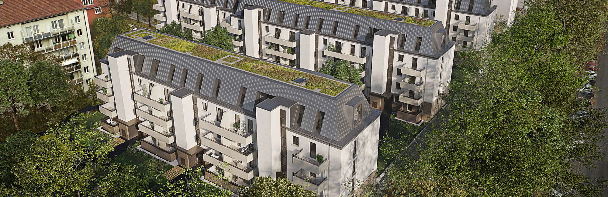 Luftaufnahme von mehreren modernen Wohngebäuden mit dunkelgrauen Mansarddächern, die teilweise begrünt sind. Die Fassaden sind hell mit hellgrauen Balkonen. Die Gebäude sind von Bäumen umgeben, und links ist ein älteres Wohnhaus zu sehen.
