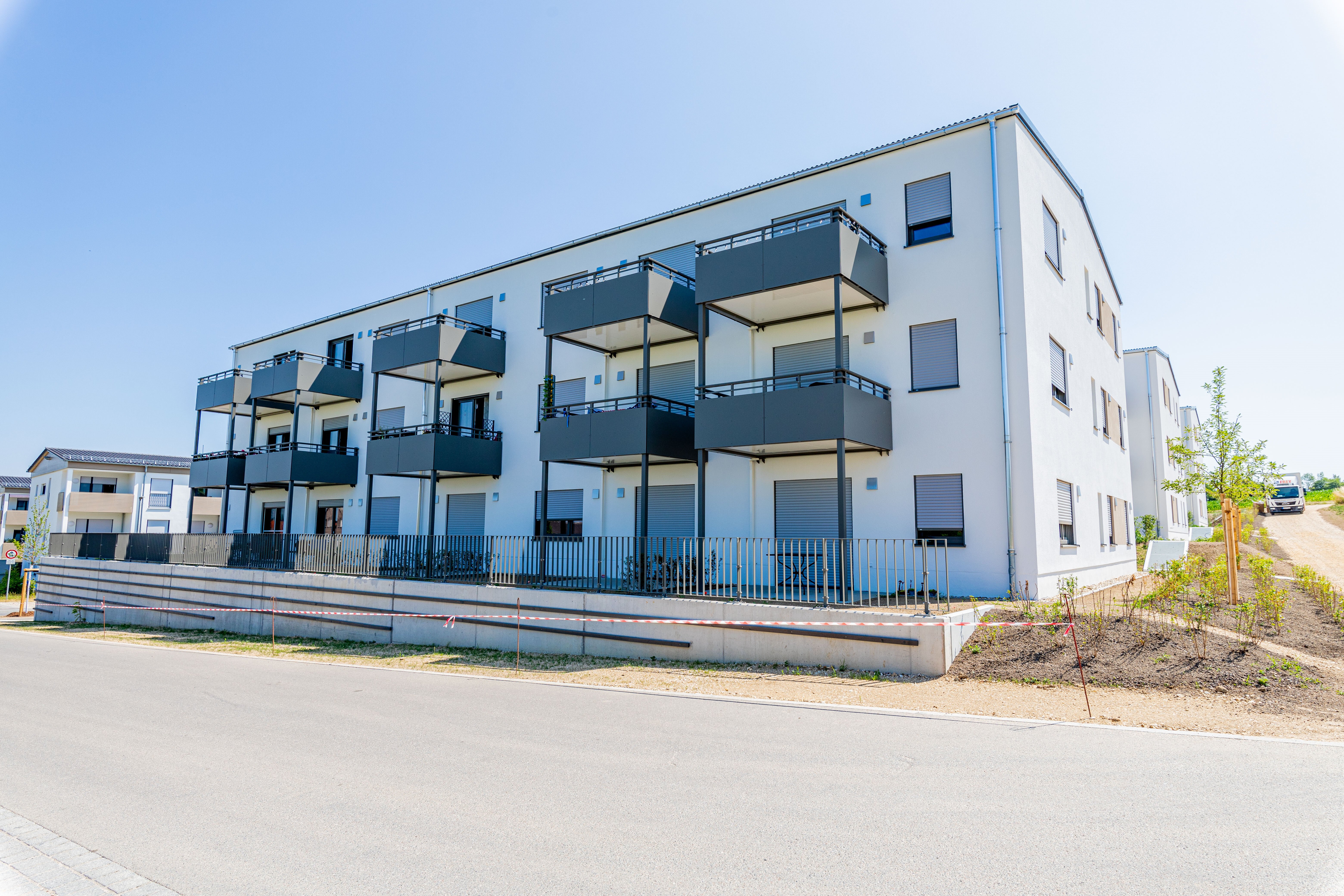 Modernes, weißes Mehrfamilienhaus mit dunklen Balkonen an einer Straße, davor ein Zaun und ein leichter Hang mit junger Bepflanzung.