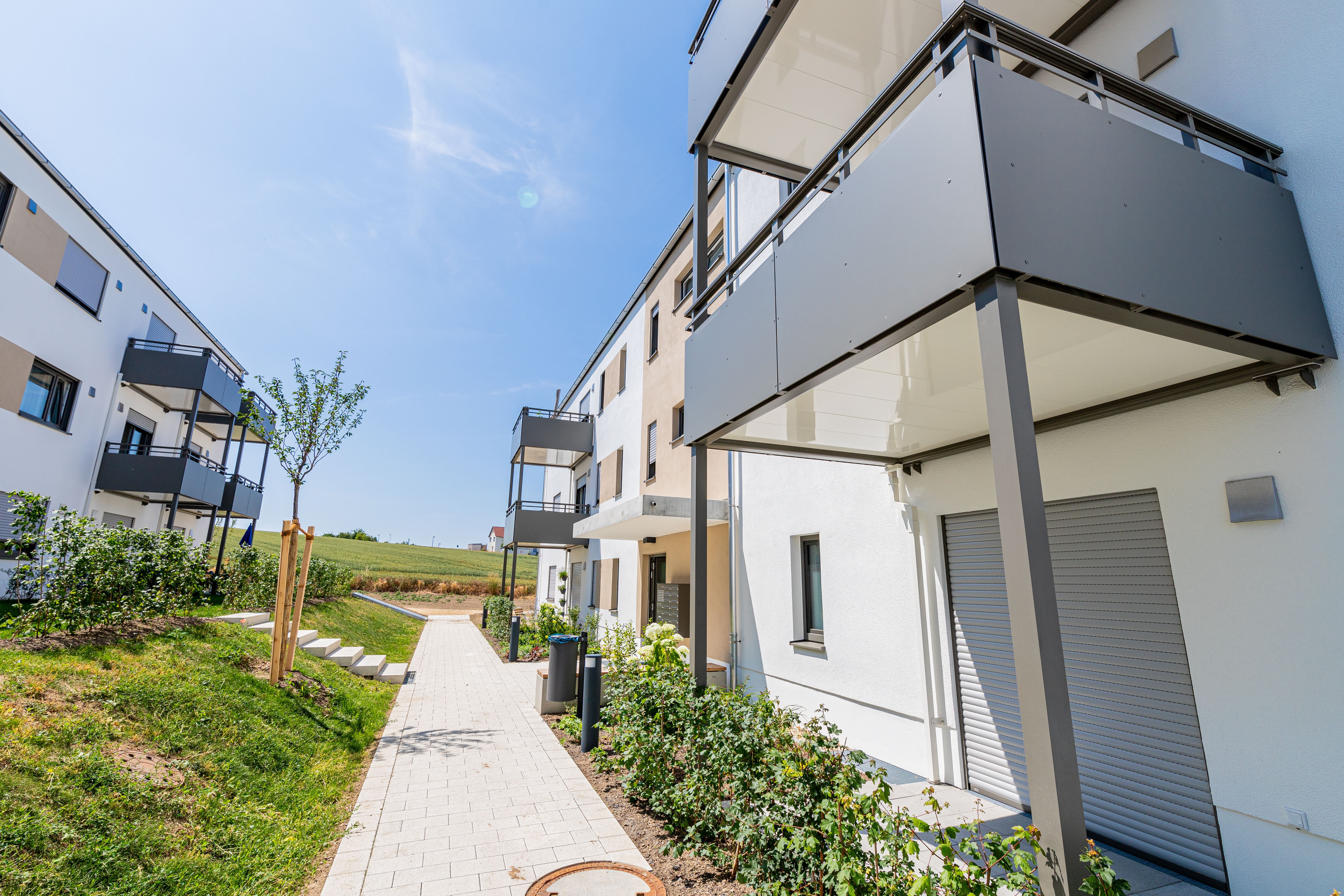 Eine gepflasterte Treppe führt zwischen zwei modernen, weißen Mehrfamilienhäusern mit dunklen Balkonen einen grünen Hang hinauf. Rechts sind die Balkone des näheren Gebäudes prominent im Bild. Im Hintergrund ist eine weite, hügelige Landschaft zu sehen.