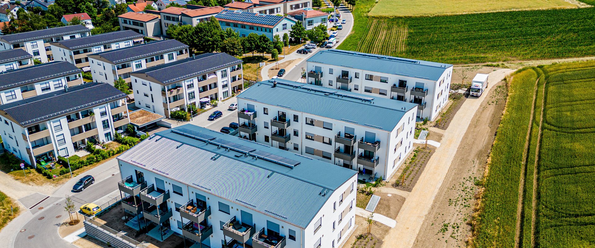  Luftaufnahme in Regensburg mit mehreren modernen, weißen Mehrfamilienhäusern mit grauen Dächern und Balkonen. Die Gebäude sind entlang einer Straße angeordnet, dahinter erstrecken sich grüne Felder und weitere Wohnhäuser in der Ferne.