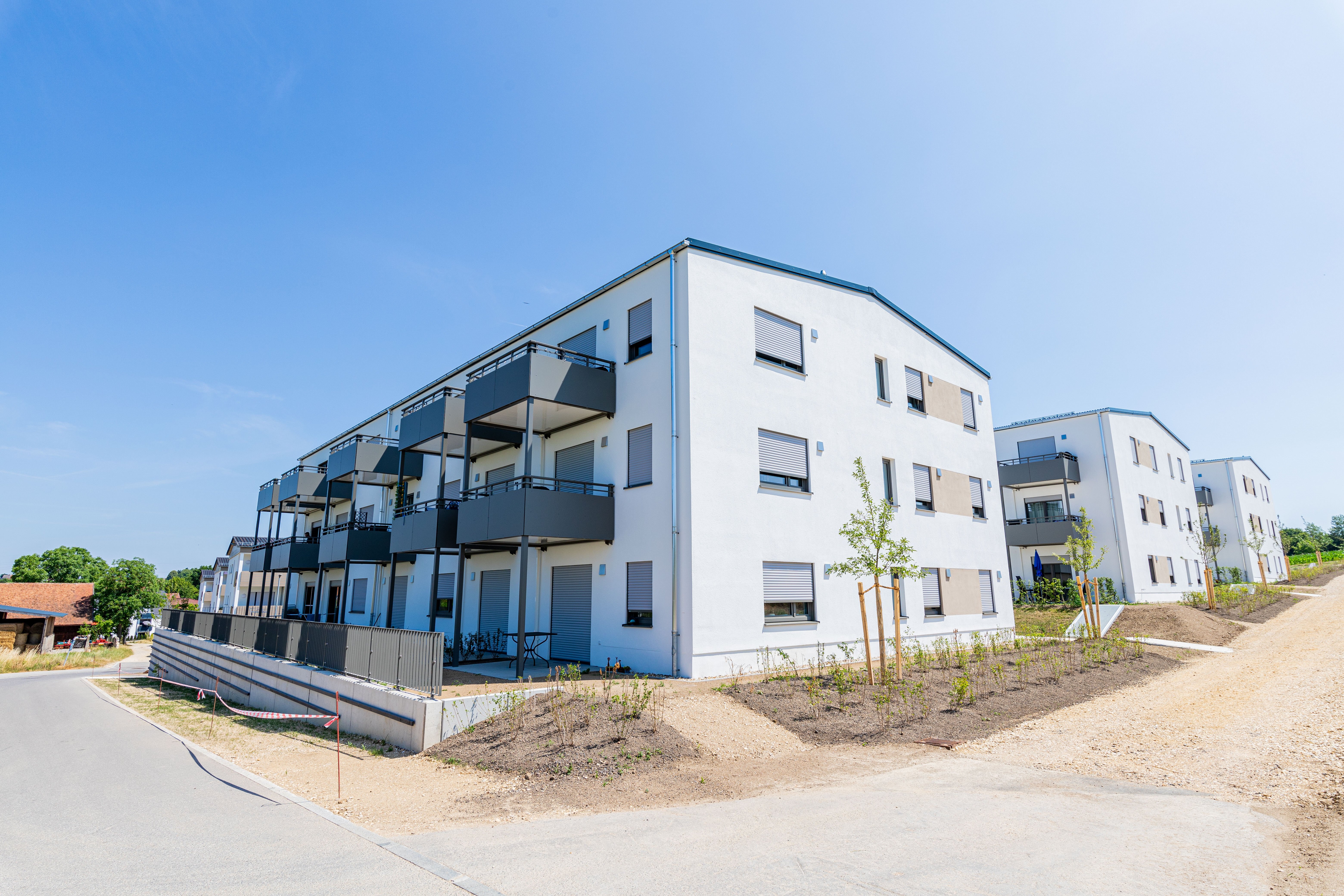  Ein modernes, weißes Mehrfamilienhaus mit dunklen Balkonen und Fensterläden steht an einer Straße. Dahinter sind weitere ähnliche Gebäude zu sehen. Im Vordergrund befindet sich ein Bereich mit Erdhügeln und junger Bepflanzung.