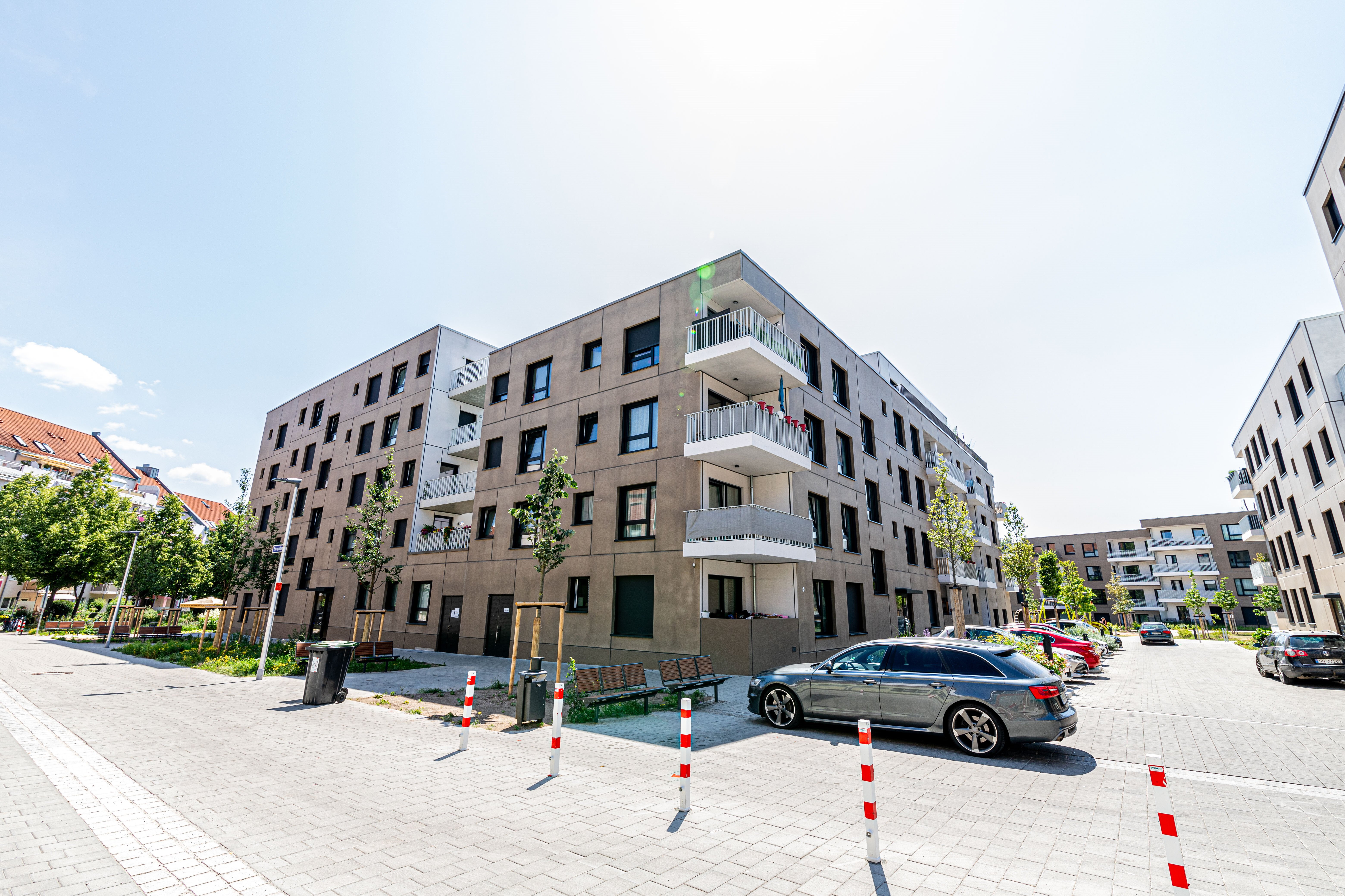 Straßenansicht einer Wohnsiedlung mit mehreren mehrstöckigen Gebäuden. Im Vordergrund steht ein Gebäude mit dunkler Fassade und hellen Balkonen. Die gepflasterte Straße ist mit rot-weiß gestreiften Pollern gesäumt und mehrere Autos sind geparkt.