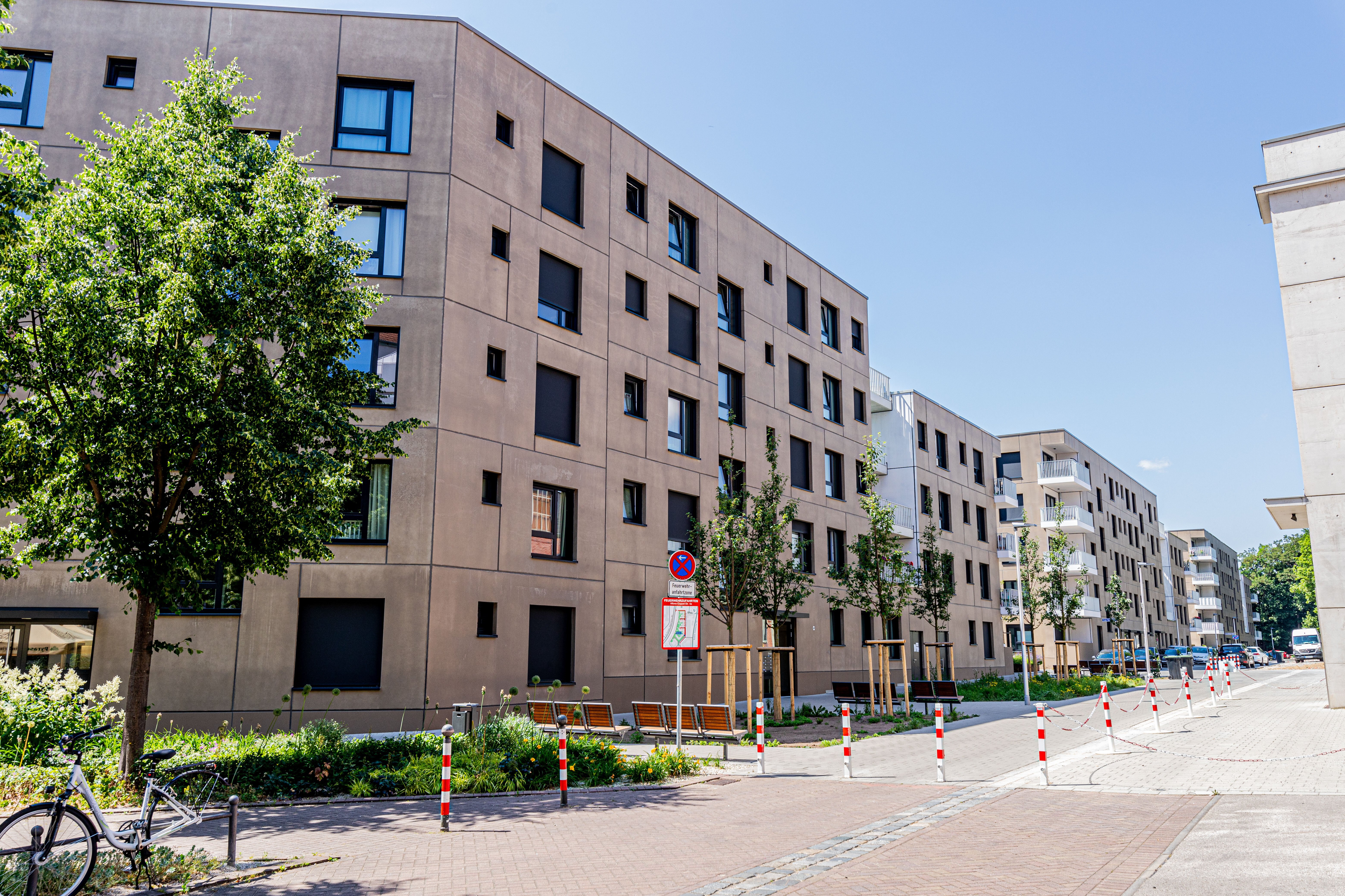 Reihe von hellen, mehrstöckigen Wohngebäuden mit vielen Fenstern und Balkonen. Im Vordergrund sind Bäume, ein Gehweg und rot-weiß gestreifte Poller zu sehen. 