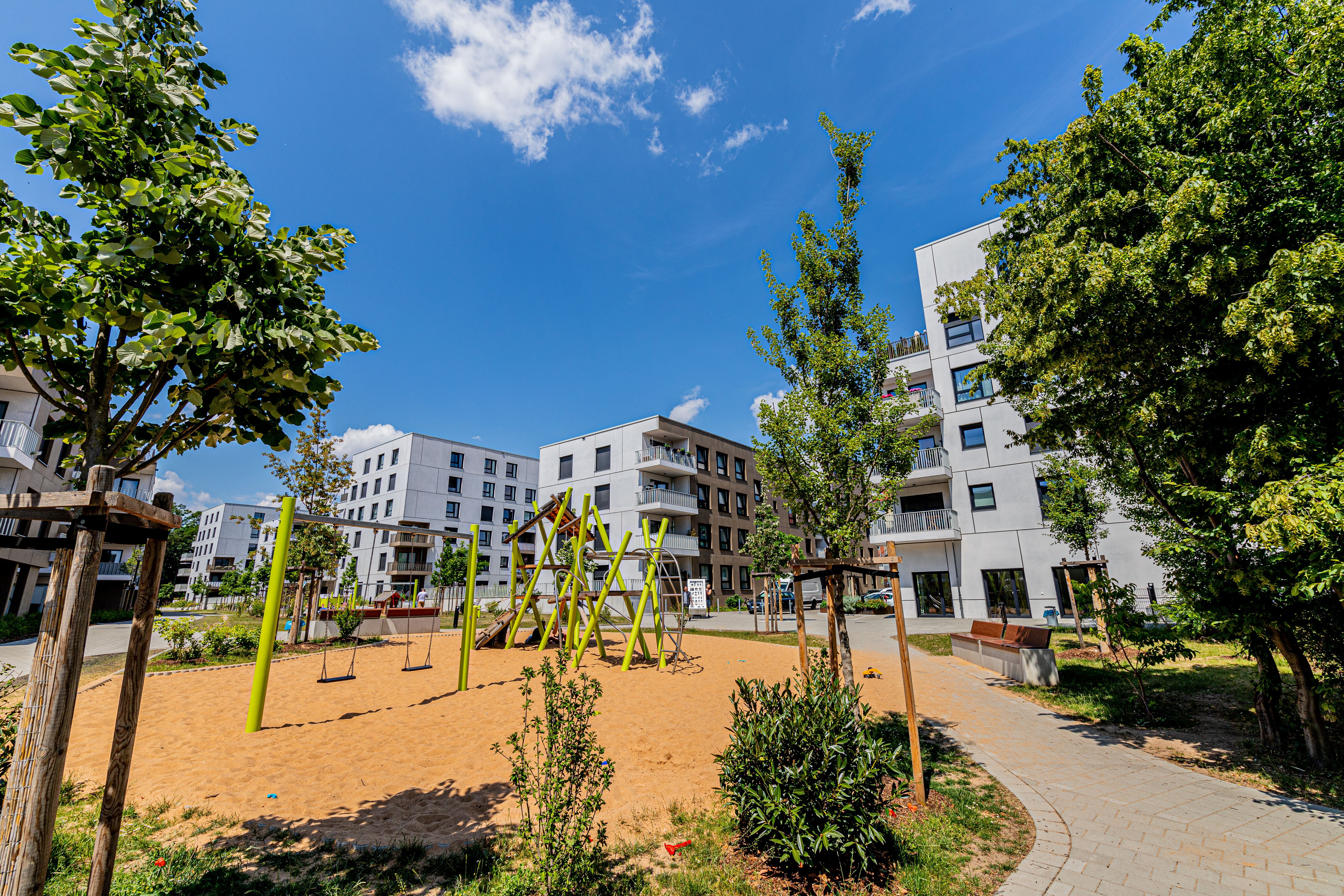 Ein Spielplatz mit Sandfläche, Schaukeln und Klettergeräten im Vordergrund. Dahinter stehen mehrere helle, mehrstöckige Wohngebäude. Bäume und ein gewundener Weg sind ebenfalls zu sehen. 