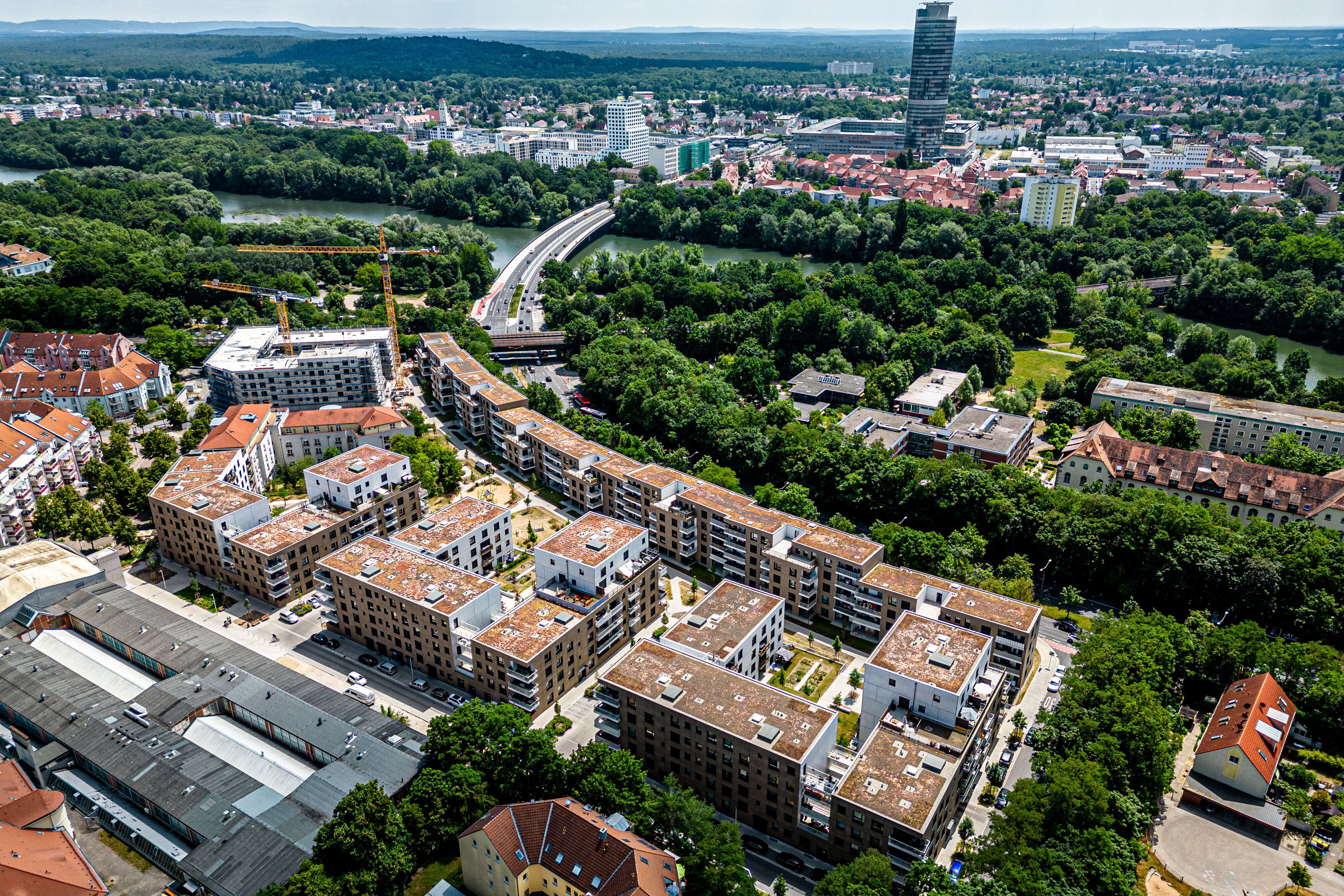 Weitläufige Luftaufnahme des Quartiers Tafel in Nürnberg, zeigt zahlreiche neue oder modernisierte Wohngebäude mit flachen Dächern, eingebettet in viel Grün. Im Hintergrund ist ein Fluss mit einer Brücke und die Skyline der Stadt mit einem Hochhaus zu sehen.