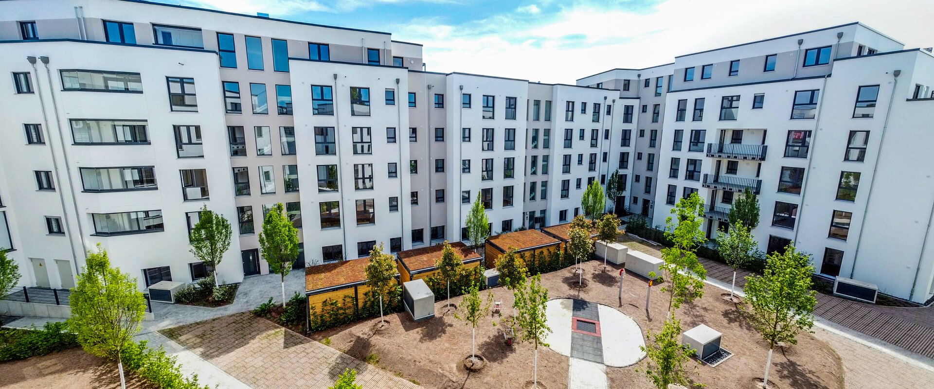  Ein moderner, weißer U-förmiger Wohnkomplex mit vielen Fenstern umschließt einen begrünten Innenhof mit Bäumen, Sitzgelegenheiten und gepflasterten Wegen. Im Vordergrund ist ein Teil eines weiteren Weges und junger Bepflanzung zu sehen. Das Datum "16.05.2022" ist unten links eingeblendet.