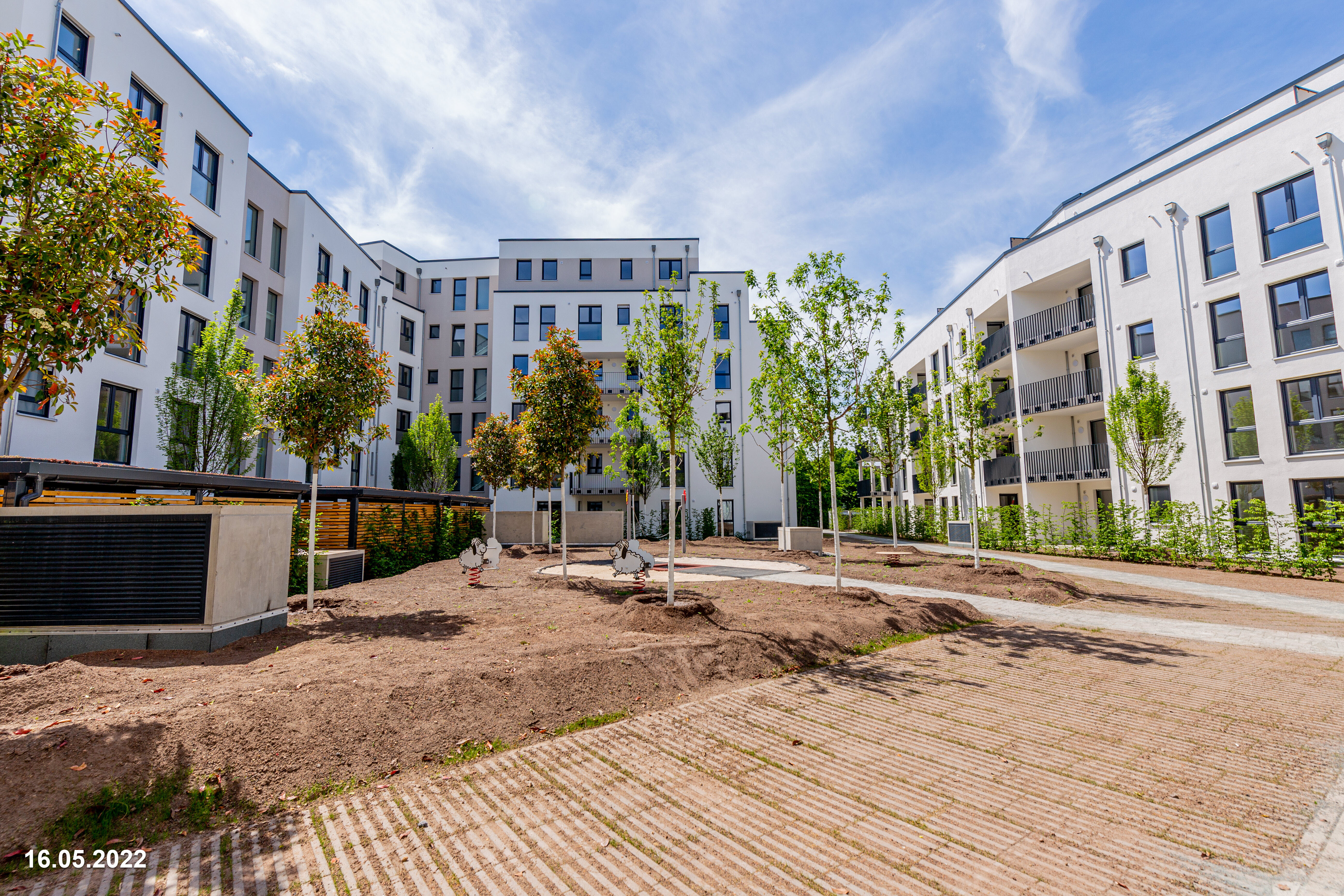  Ein weitläufiger Außenbereich mit unbebauten Erdbereichen und jungen Bäumen zwischen modernen, weißen Wohngebäuden. Rechts ist ein gepflasterter Weg zu sehen. Das Datum "16.05.2022" ist unten links eingeblendet.