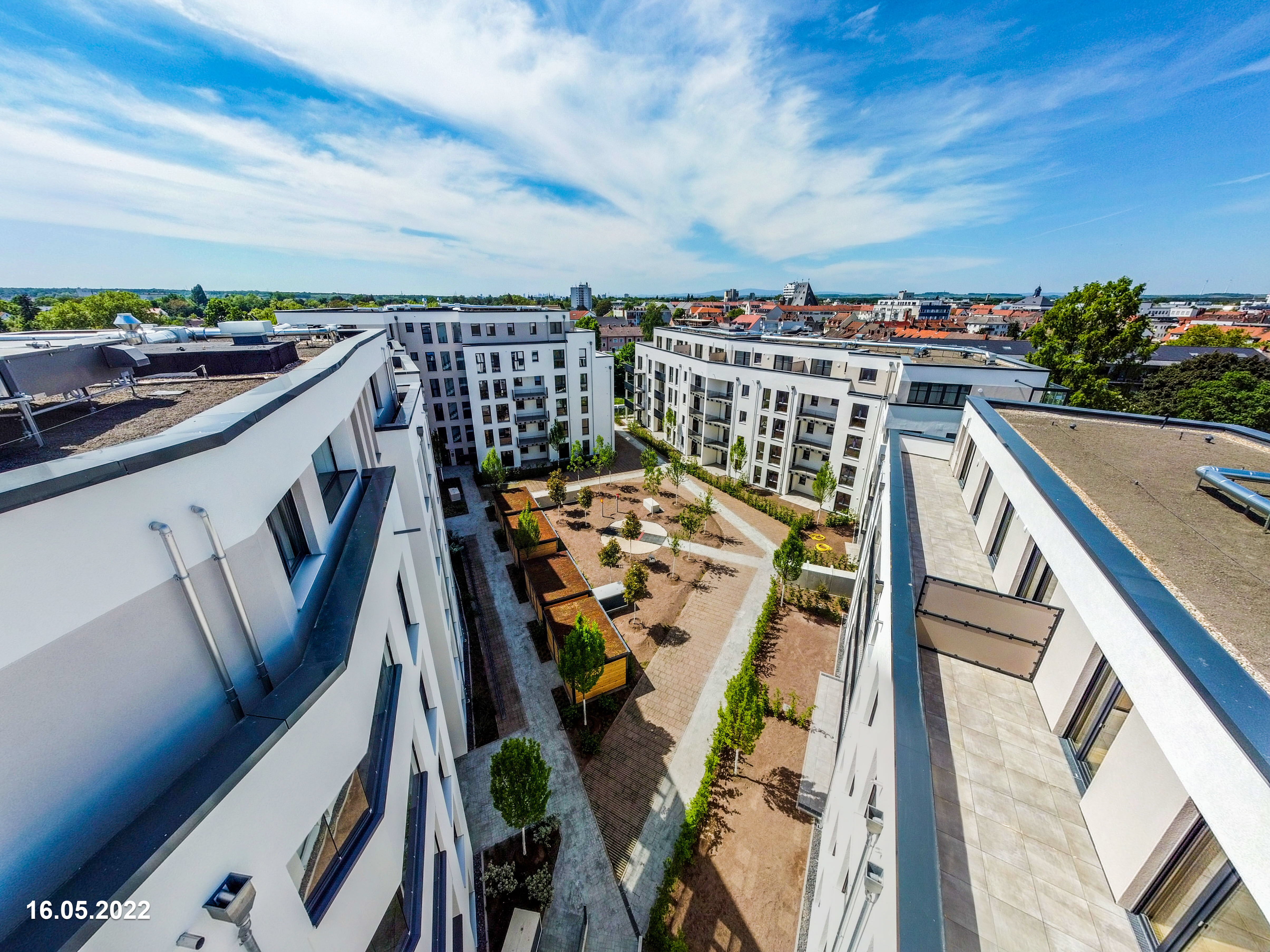  Luftaufnahme eines modernen Wohnkomplexes mit weißen Gebäuden, die einen begrünten Innenhof umschließen. Von oben blickt man auf die Flachdächer der Gebäude, dahinter erstreckt sich eine Stadtlandschaft mit weiteren Gebäuden und Bäumen unter einem weiten Himmel. Das Datum "16.05.2022" ist unten links eingeblendet.