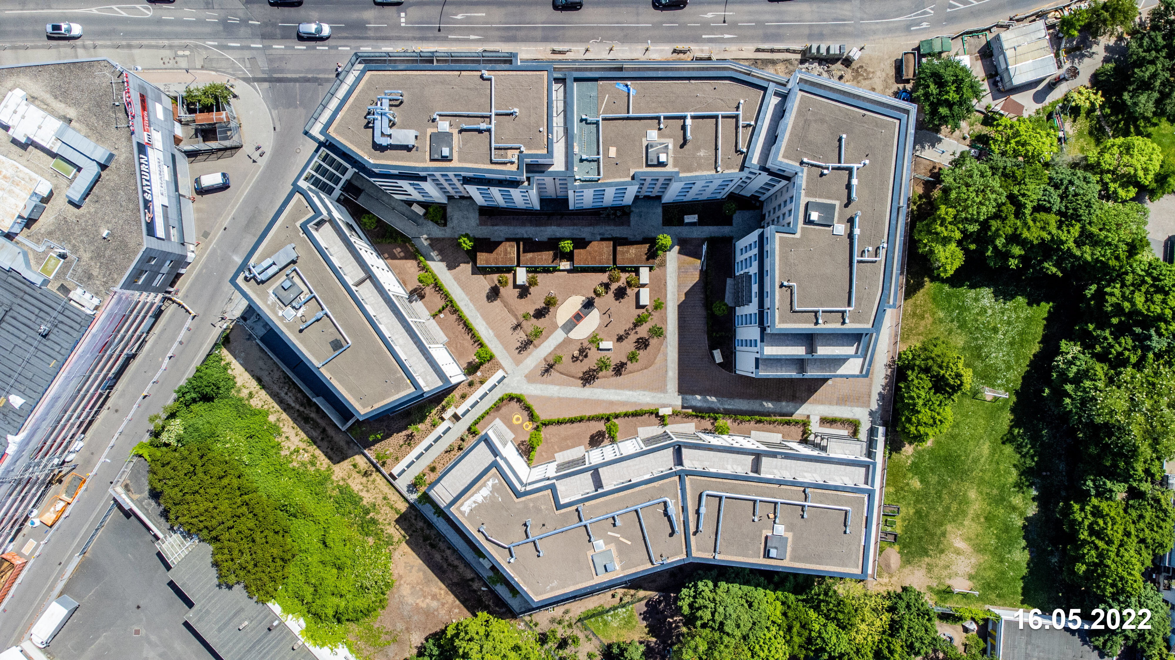 Luftaufnahme eines modernen Gebäudekomplexes mit mehreren Flügeln, die einen zentralen, begrünten Innenhof umschließen. Die Dächer sind flach und grau, die Fassaden weiß. Das Gebäude ist von Straßen und Grünflächen umgeben. Das Datum "16.05.2022" ist unten links und rechts eingeblendet.