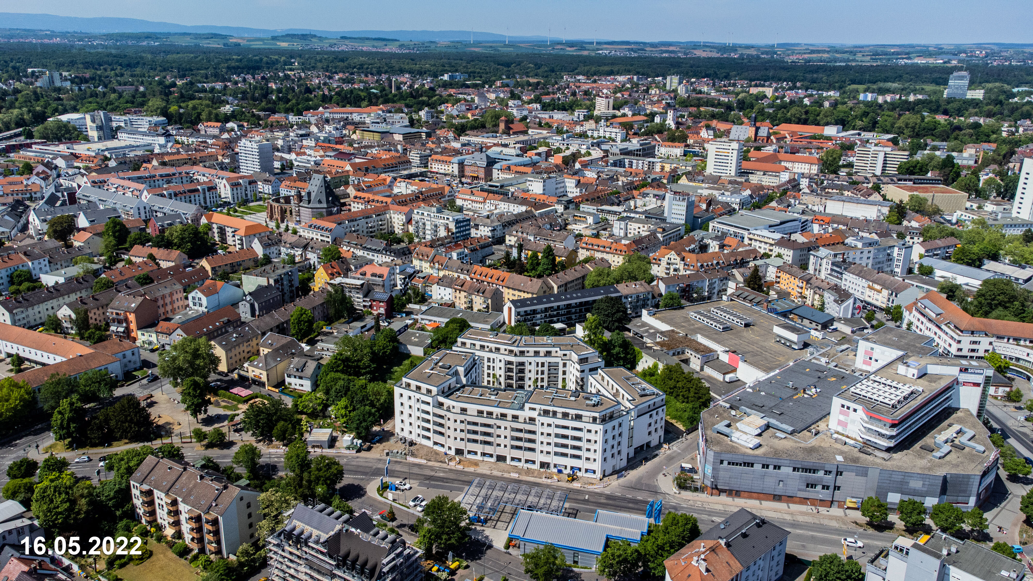 Luftaufnahme einer weitläufigen Stadtlandschaft mit vielen Gebäuden unterschiedlicher Größe und Dächerfarben, darunter ein großer, weißer Gebäudekomplex im Vordergrund. Die Stadt ist von Grünflächen und Wäldern umgeben. Das Datum "16.05.2022" ist unten links eingeblendet.
