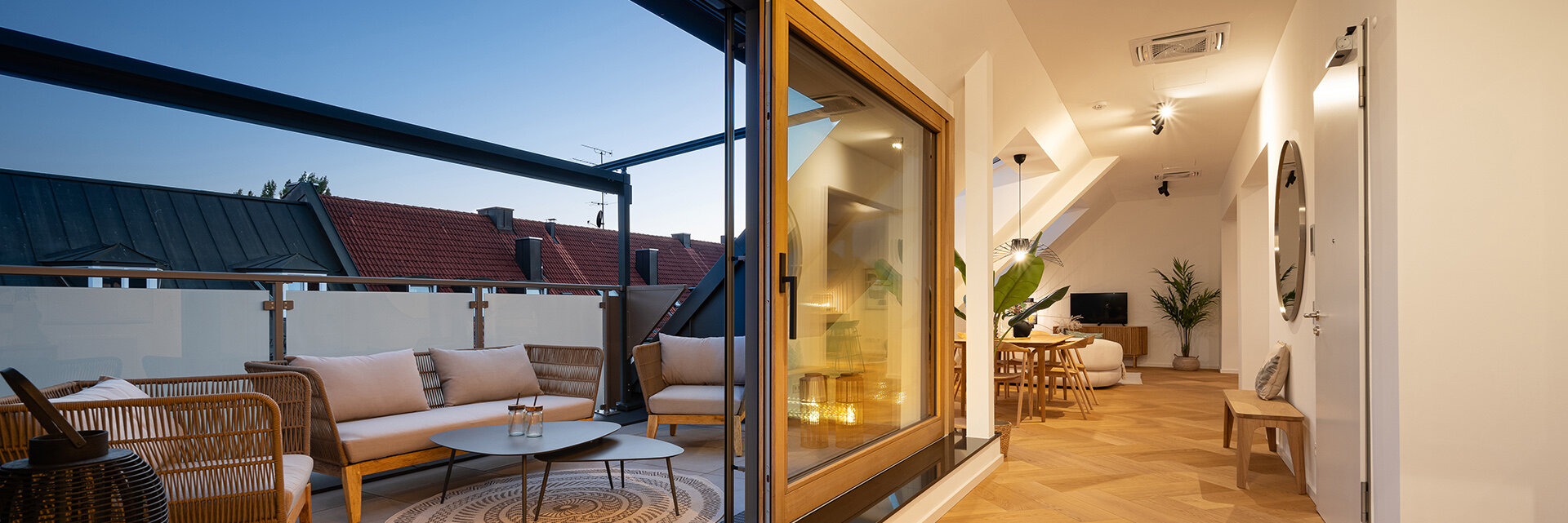 Wohnraum und Balkonanblick der Dachgeschosswohnung des Kaufobjekts Bohome. Beleuchtet und gemütlich eingerichtet mit Balkonmöbeln, Esstisch, Sofa, Fernseher und Pflanzen