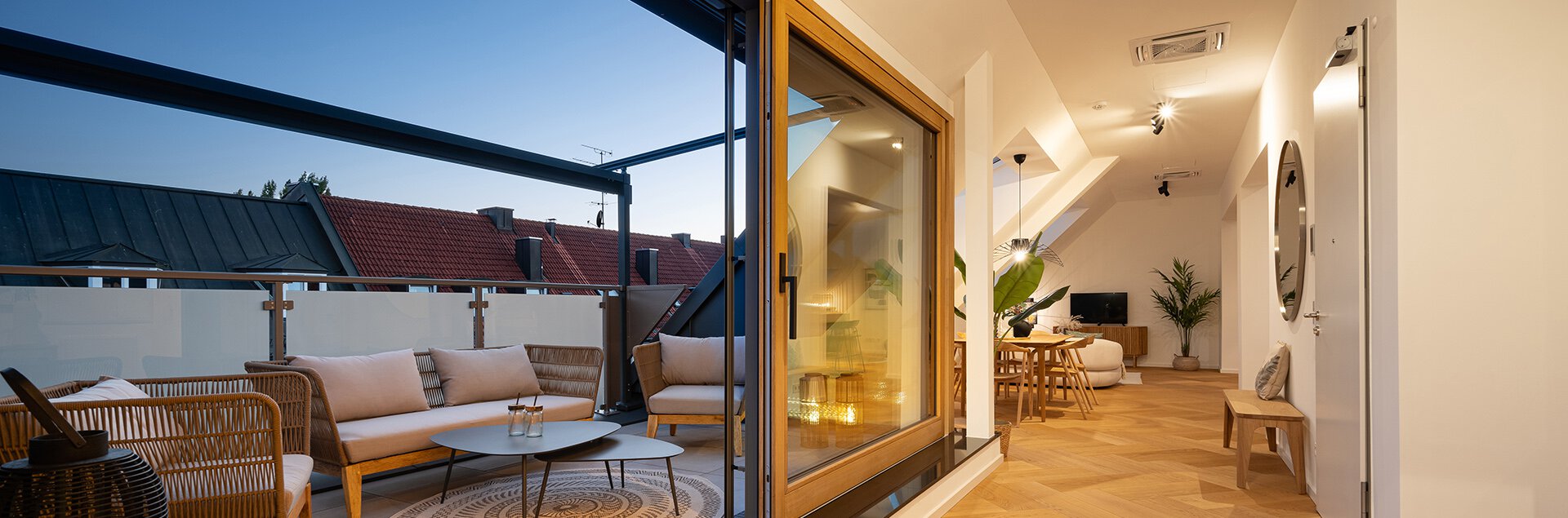 Wohnraum und Balkonanblick der Dachgeschosswohnung des Kaufobjekts Bohome. Beleuchtet und gemütlich eingerichtet mit Balkonmöbeln, Esstisch, Sofa, Fernseher und Pflanzen
