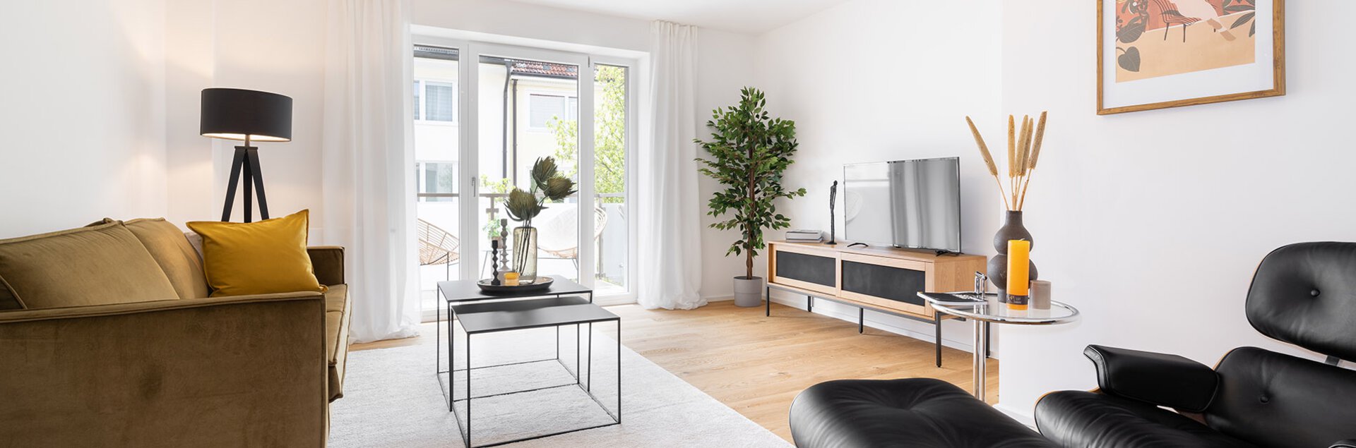 Modernes, helles Wohnzimmer mit senfgelbem Samtsofa, schwarzem Design-Sessel, Holzboden und einer großen Fensterfront zum Balkon.