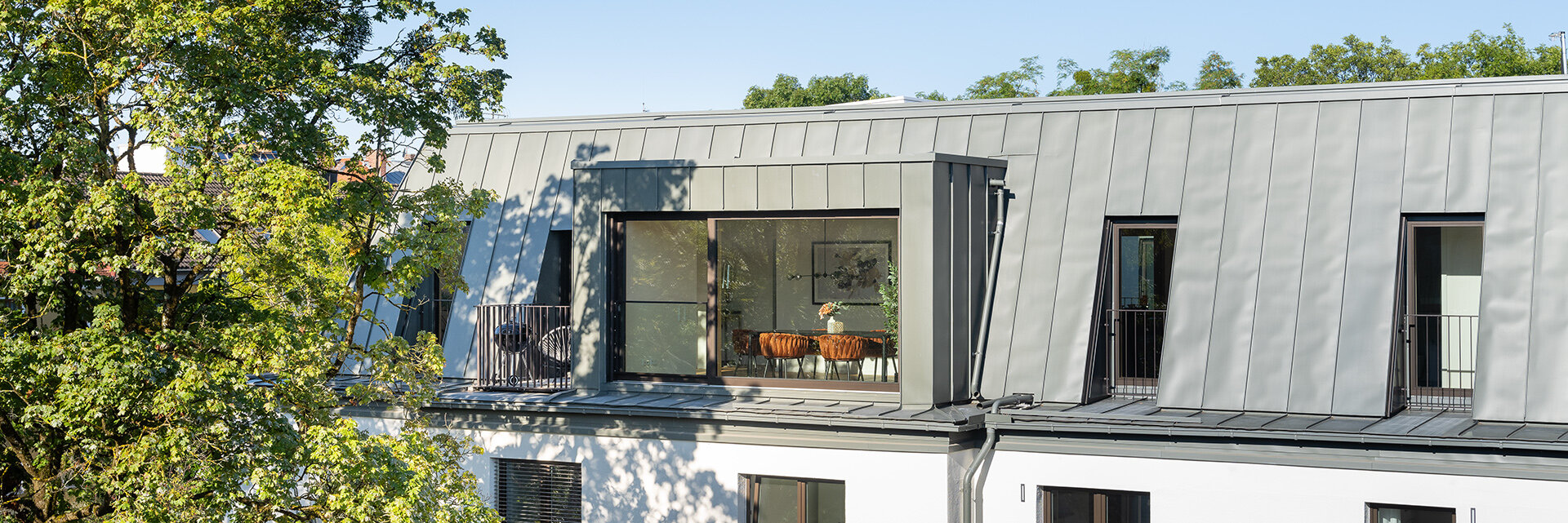 Blick auf Noya Dachgeschosswohnung aus gleicher Höhe. Blick auf den Balkon und durch die große Fensterfront auf den Esszimmertisch. Ein großer Baum vor dem Wohngebäude spendet Schatten auf dem Balkon