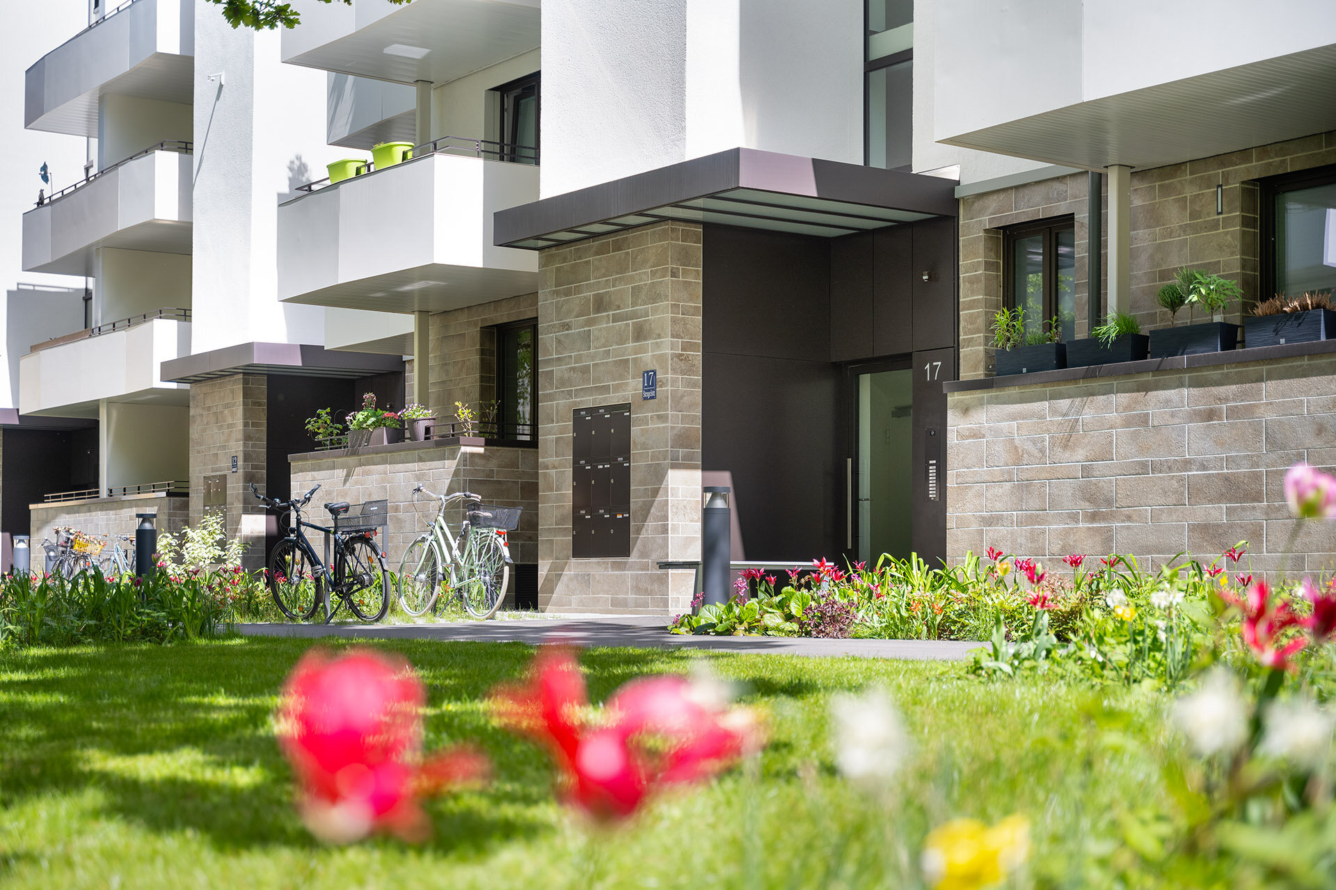 Blick von der Wiese und Blumenbeeten auf den Eingangsbereich und die Erdgeschosswohnungen des modernen Wohngebäudes. Steinvertäfelung der Erdgeschosswohnung mit Balkonen 