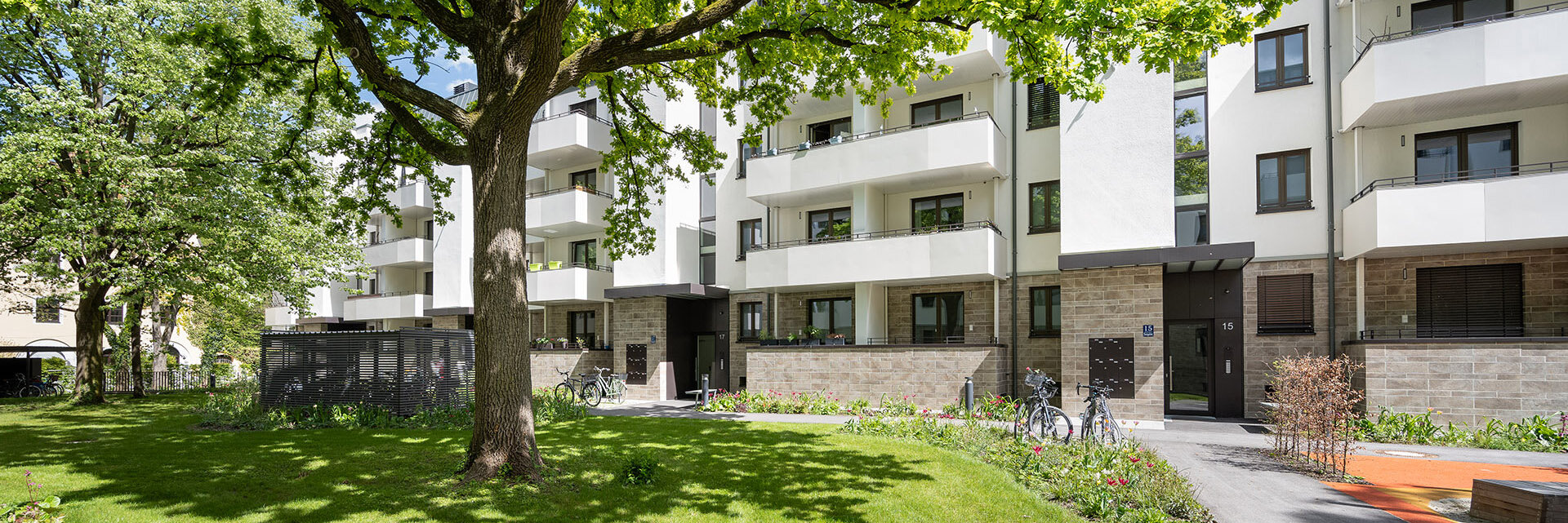Blick von grüner Wiese auf modernes Wohngebäude mit weißer Fassade, Erdgeschoss mit Steinvertäfelung und geräumigen Balkonen auf jeder Etage. 