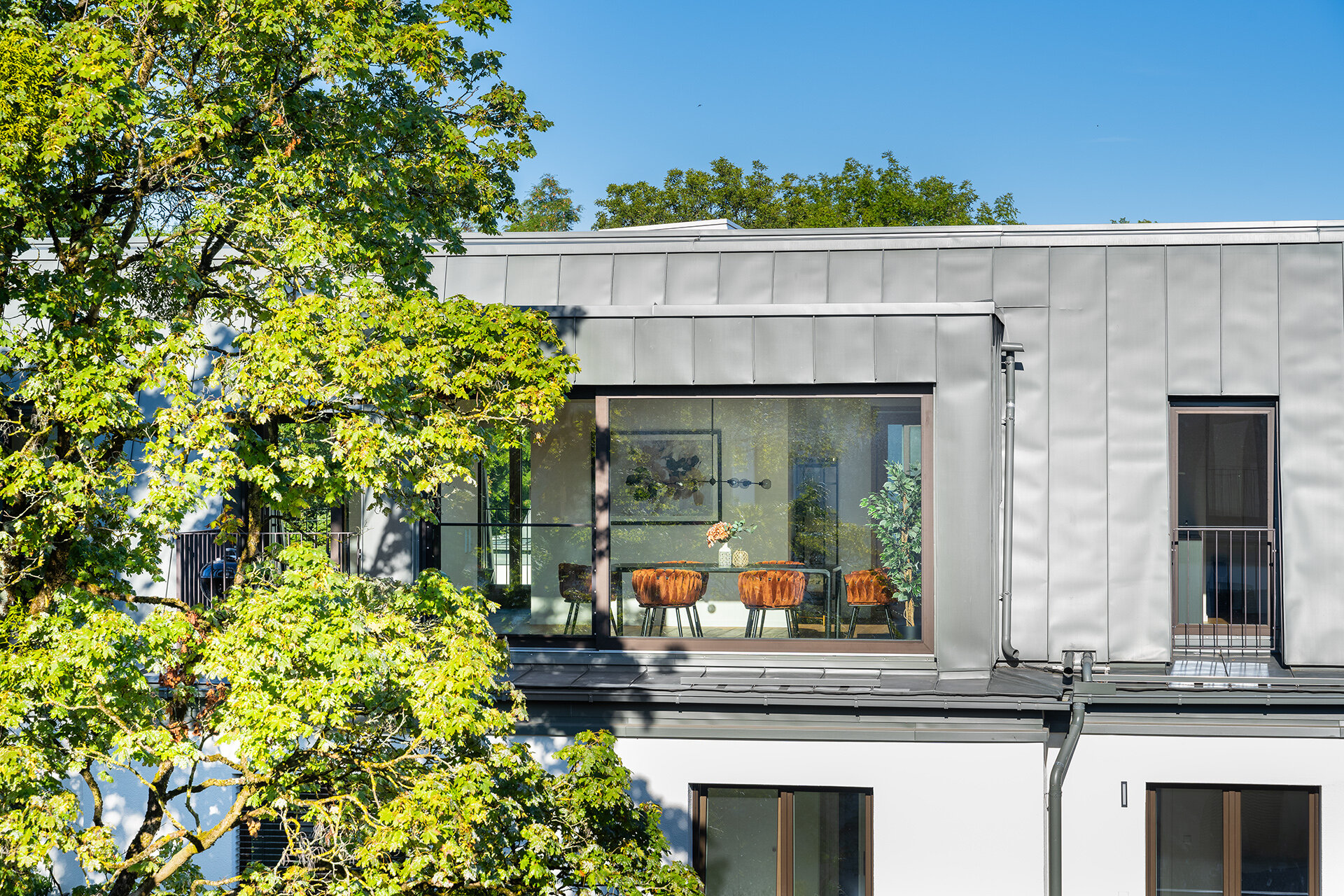 Blick auf Neubau Dachgeschosswohnung durch die große Fensterfront auf den Esszimmertisch. Ein hoher Baum neben dem Gebäude spendet Schatten auf dem angrenzenden Balkon