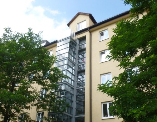 Straßenansicht des Kaufobjekts in der Morawitzkystraße mit Außenaufzug und umgebenden Bäumen.