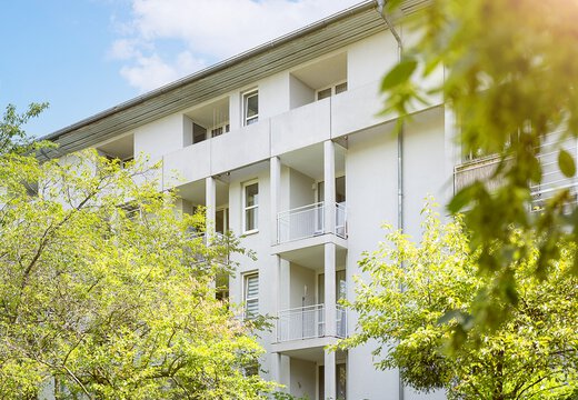 Blick auf das Wohngebäude mit weißer Fassade und Balkonen auf jeder Etage umgeben von hohen Laubbäumen 