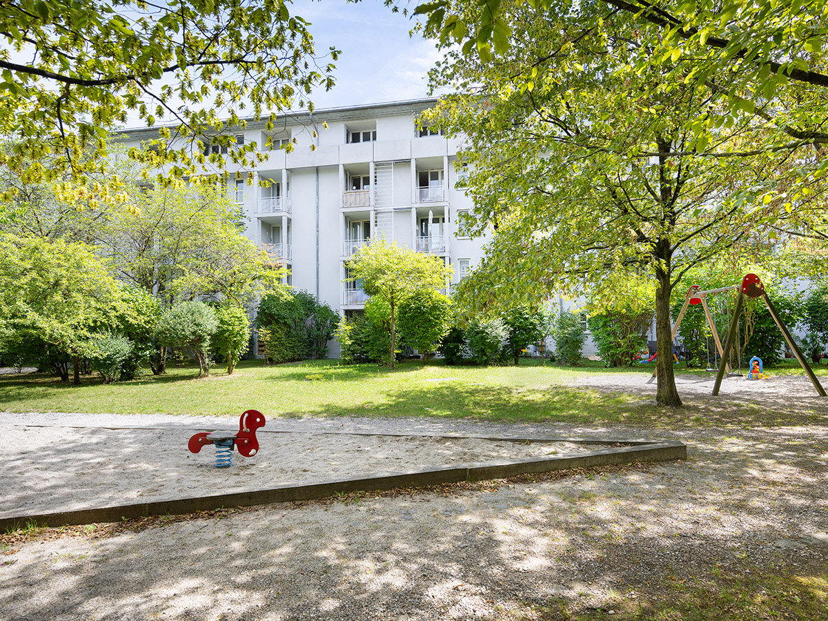 Rückseite des Wohnkomplex mit Garten, hohen Laubbäumen und Spielplatz. Blick vom Balkon in die Grünanlage