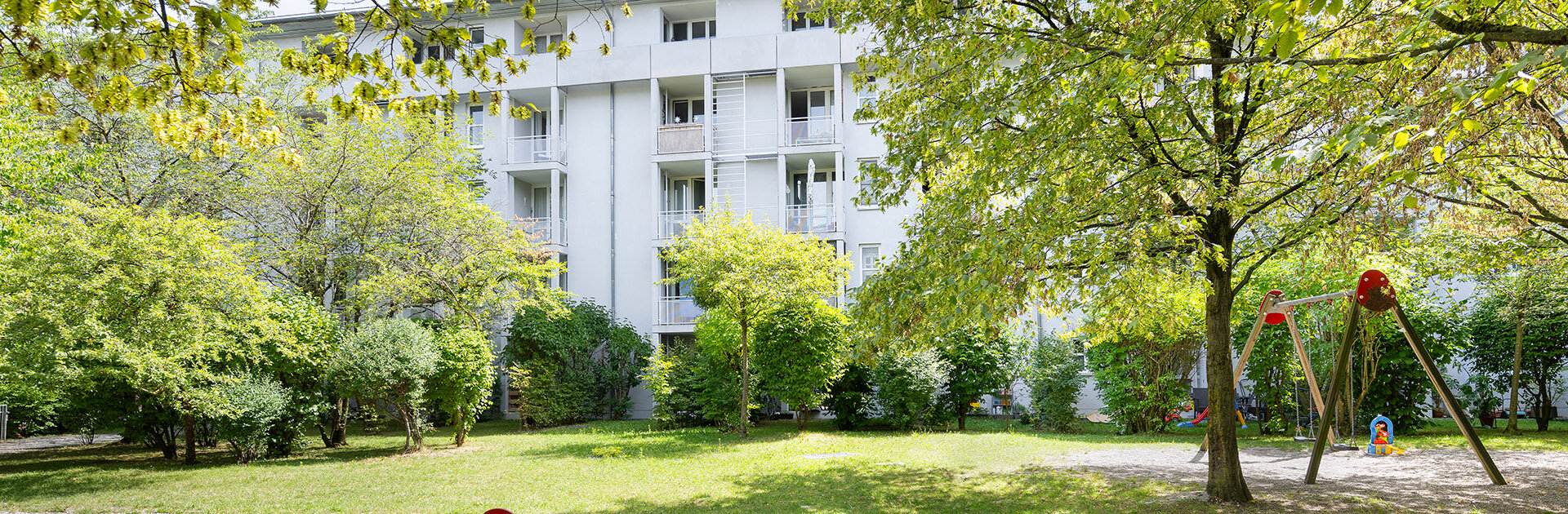 Rückseite des Wohnkomplex mit Garten, hohen Laubbäumen und Spielplatz. Blick vom Balkon in die Grünanlage