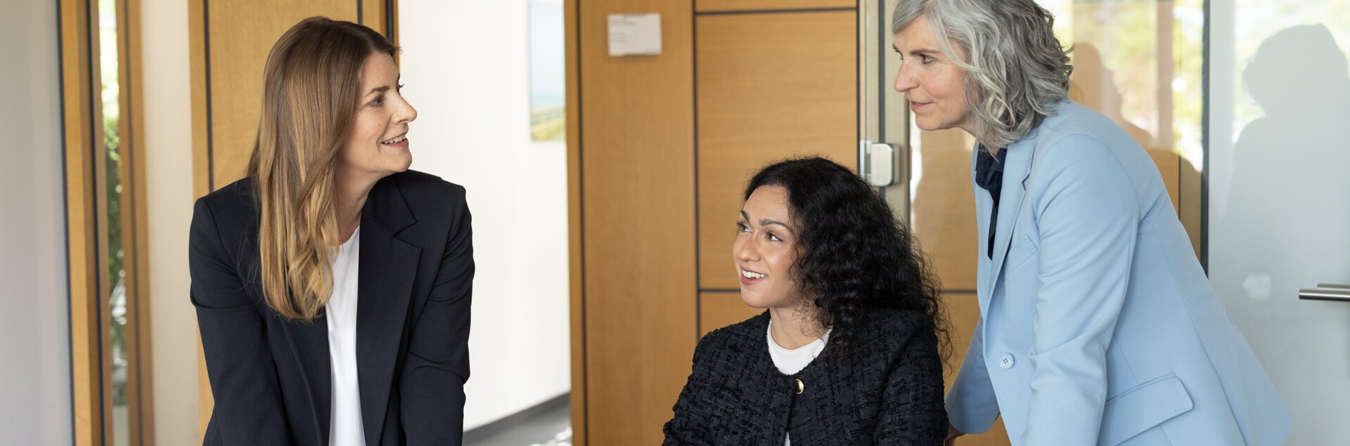 Drei Frauen in Business-Kleidung unterhalten sich lächelnd in einem Büro.