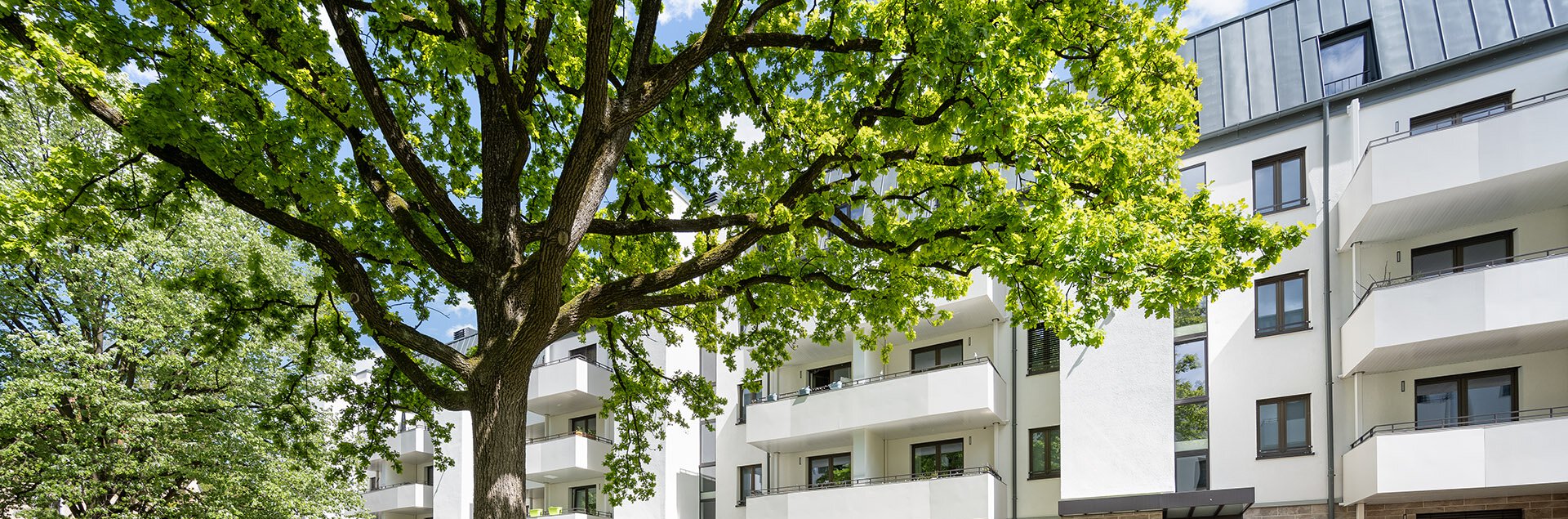 Modernes Wohnhaus mit Balkonen, umgeben von grüner Rasenfläche, Bäumen und einem Zugangsweg.
