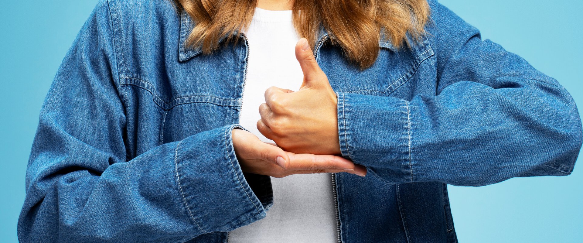 Person in blauer Jeansjacke vor einfarbig blauem Hintergrund zeigt mit beiden Händen das Gebärdenzeichen für 'Ich liebe dich'.