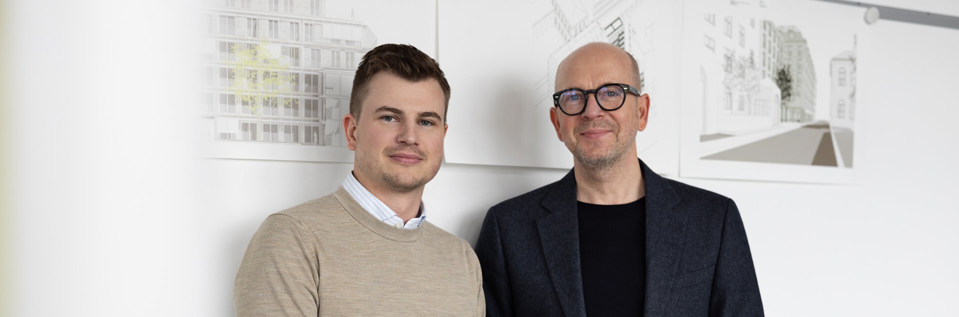 Zwei Männer in Business-Kleidung stehen lächelnd in einem Büro vor Bauplänen.