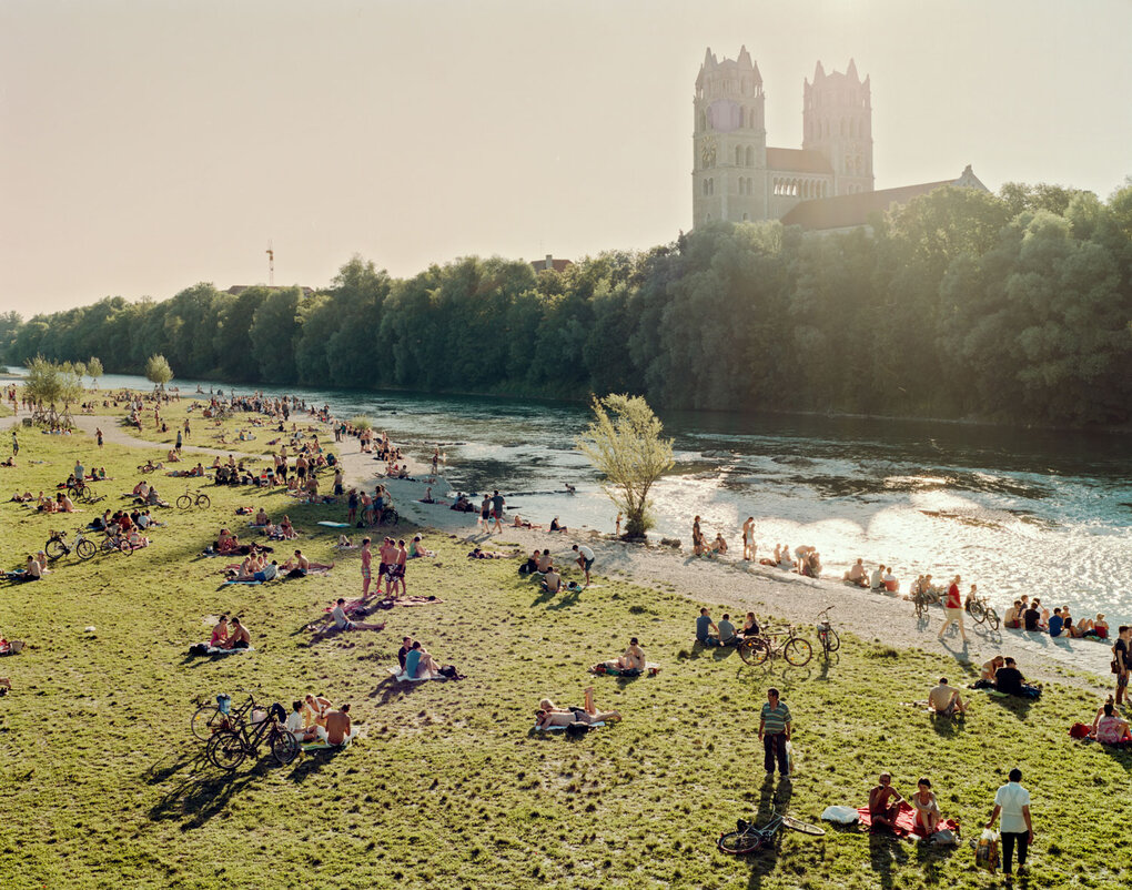 Isarufer in München an einem sonnigen Tag. Viele Menschen entspannen am Flussufer mit Blick auf die Frauenkirche.
